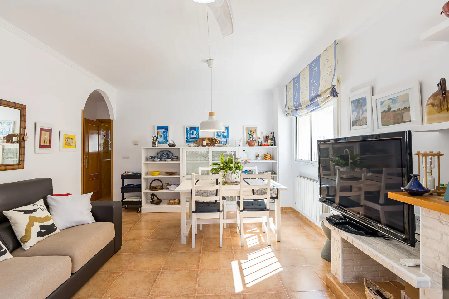 Bright living room with a brown sofa, white dining table and chairs, and a large TV. Shelves display decorative items. Tiled floor.