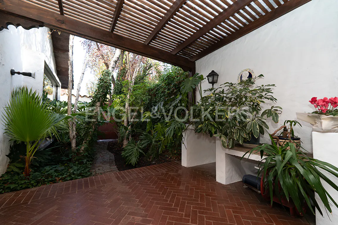 Outdoor patio with brick flooring, a wooden pergola, and lush greenery. White walls with plants and a garden path are visible.
