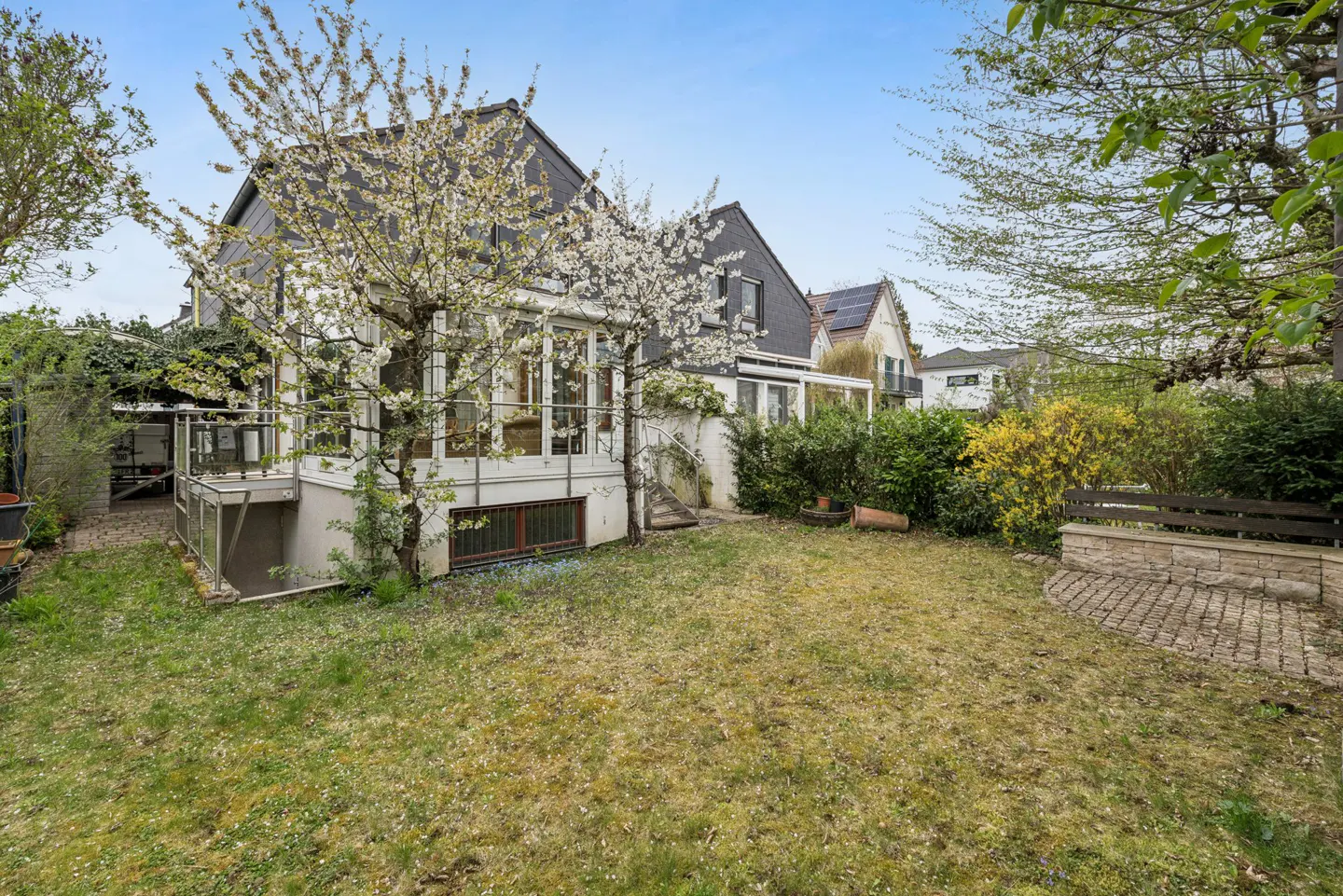 Backyard view of a two-story house with a gray roof, white walls, and a glass-enclosed porch, surrounded by green grass and trees.