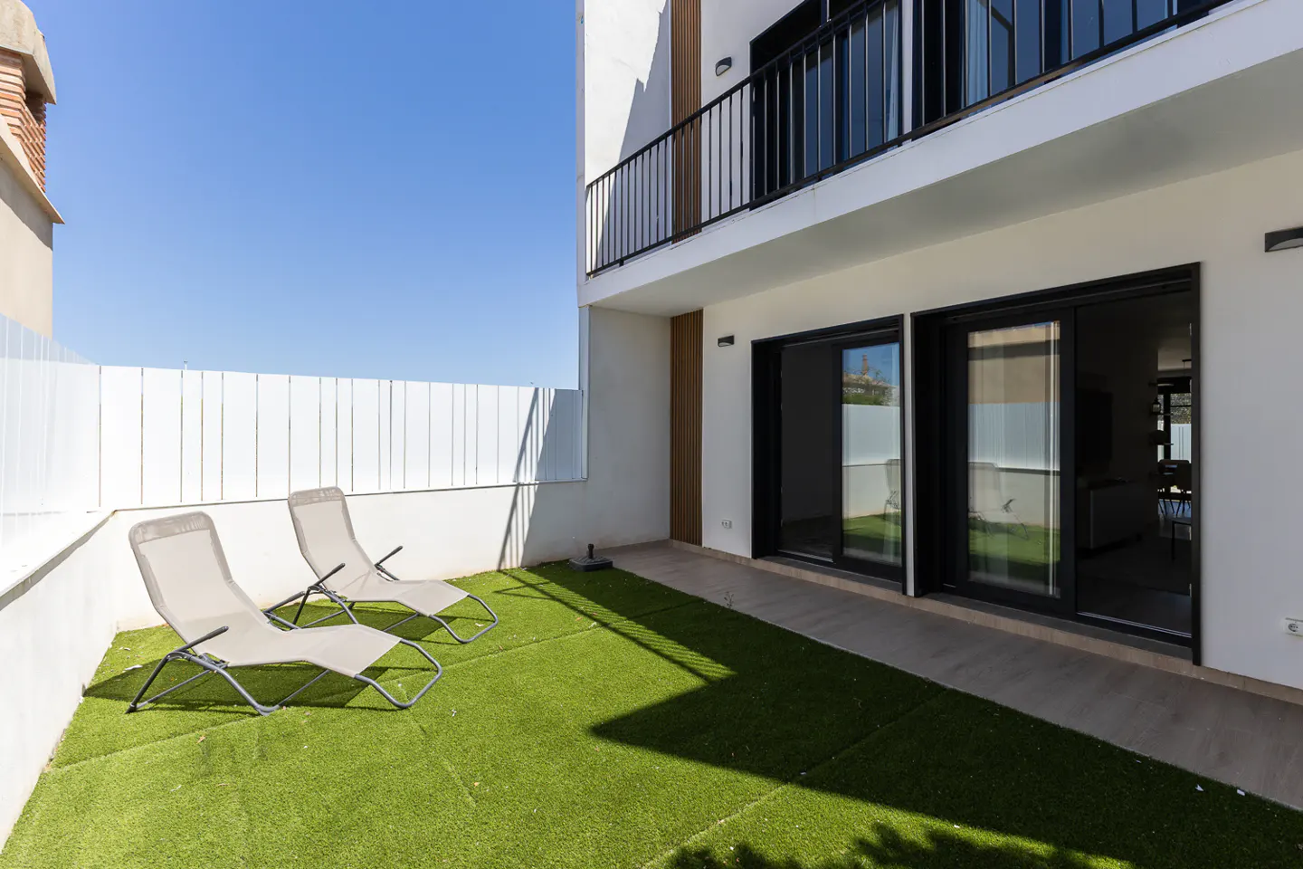 A modern home's backyard with two lounge chairs on green turf, a white fence, and sliding glass doors leading inside.