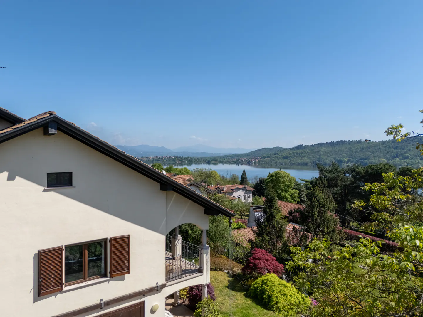 View of a cream-colored house with brown shutters and a balcony, overlooking a lake surrounded by green trees under a clear blue sky.