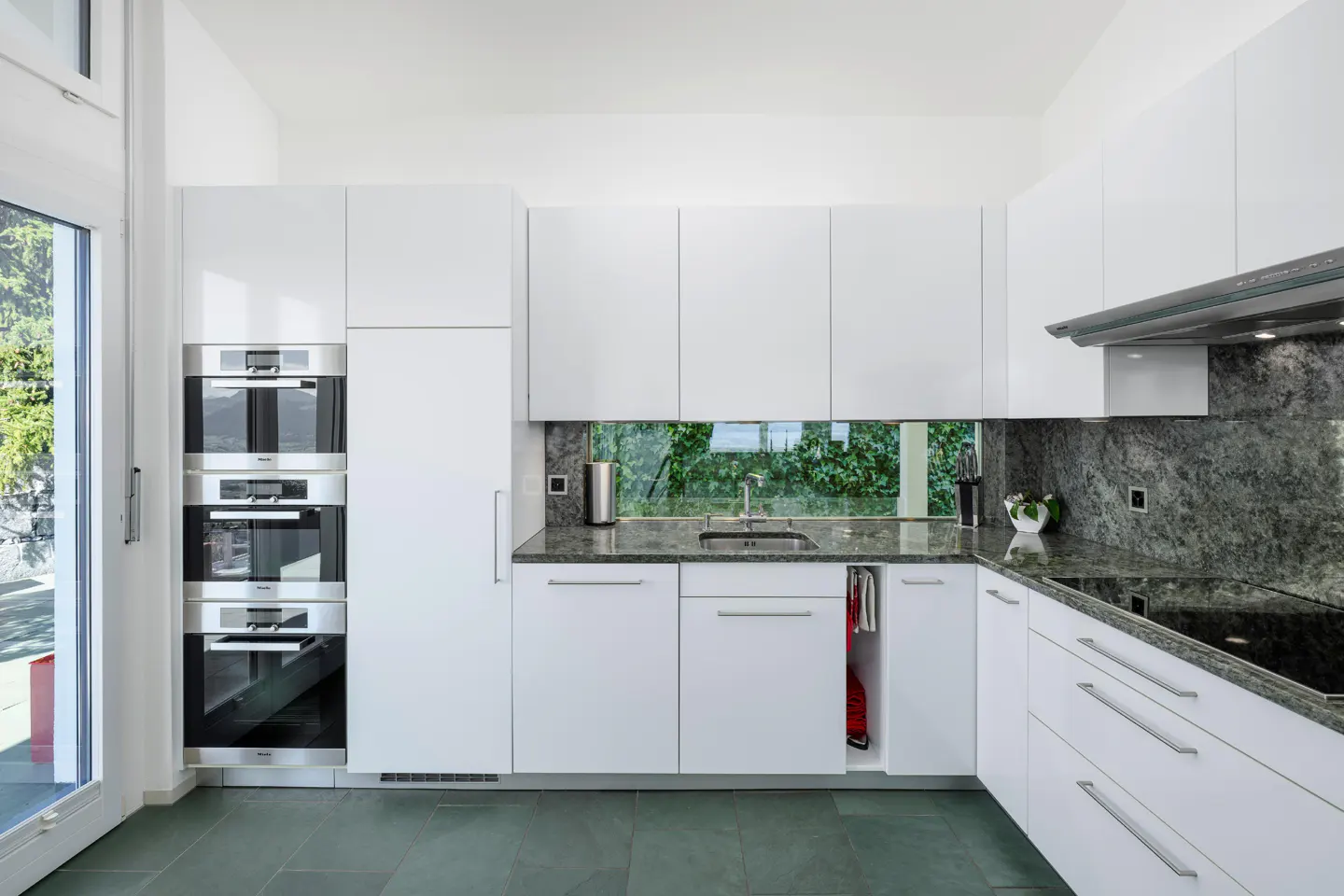 A modern kitchen with white cabinets, gray granite countertops, and stainless steel appliances. A window above the sink offers a view of greenery.
