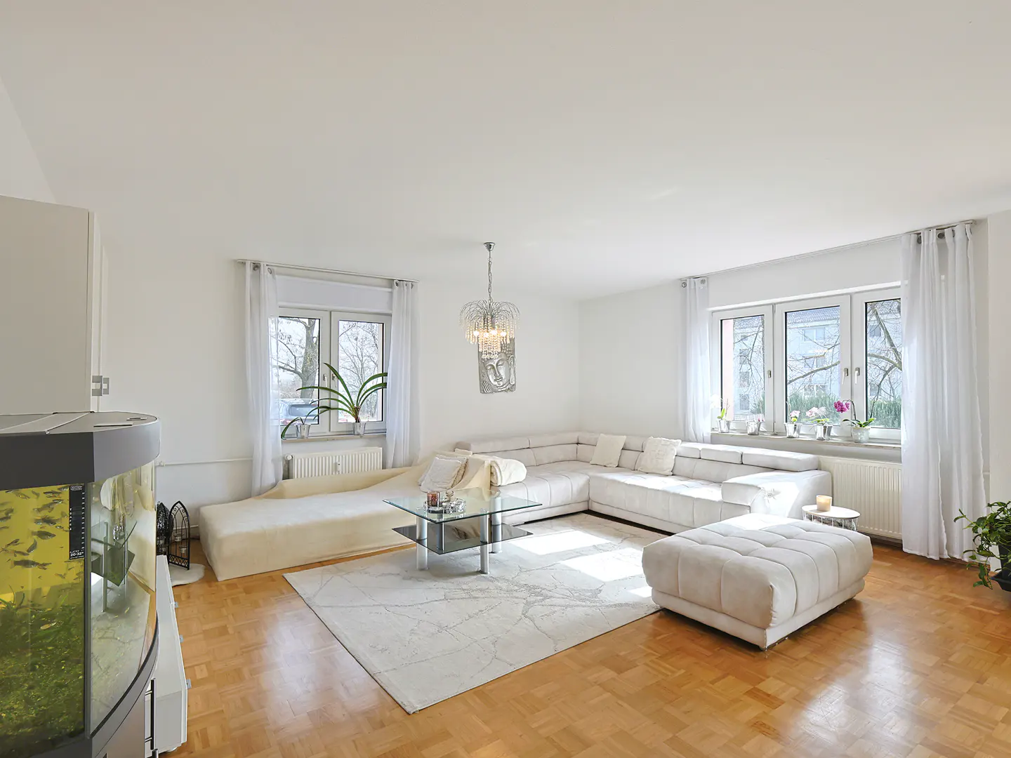 Bright living room with white sectional sofa, ottoman, glass table, and light wood floors. Windows with sheer curtains let in natural light.