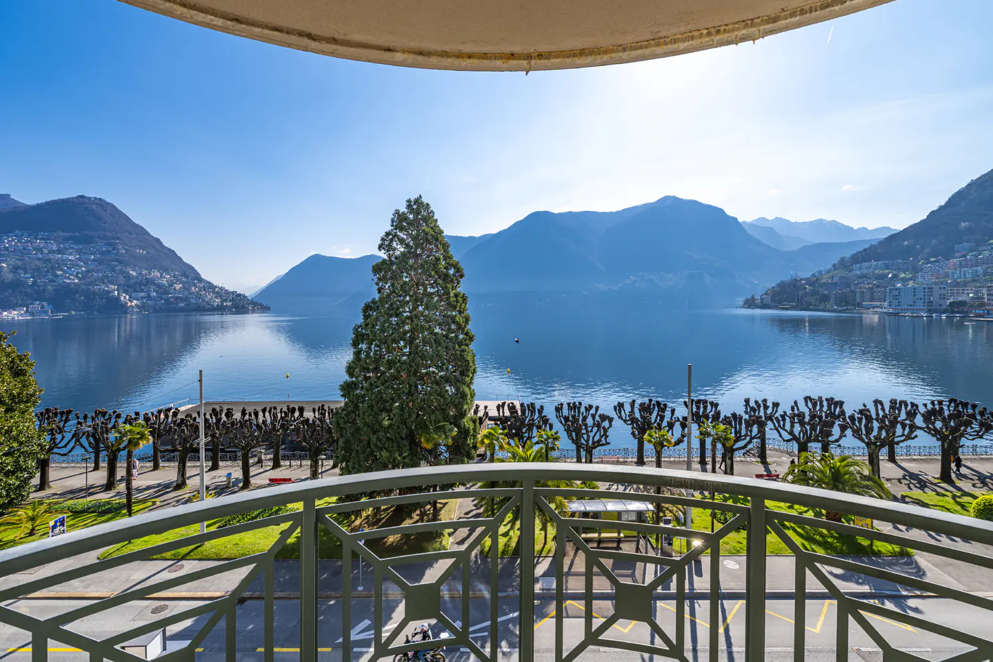 View from a balcony overlooking a blue lake, mountains, and a tree-lined promenade on a sunny day.