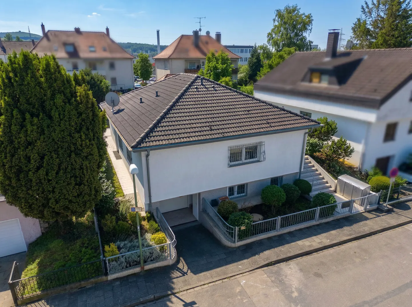 Aerial view of a two-story white house with a brown tiled roof, surrounded by green trees and bushes. A metal fence encloses the front yard.