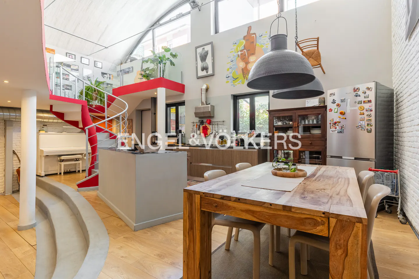 Open-concept loft with a wooden dining table, kitchen island, stainless steel fridge, and a red spiral staircase leading to a mezzanine.
