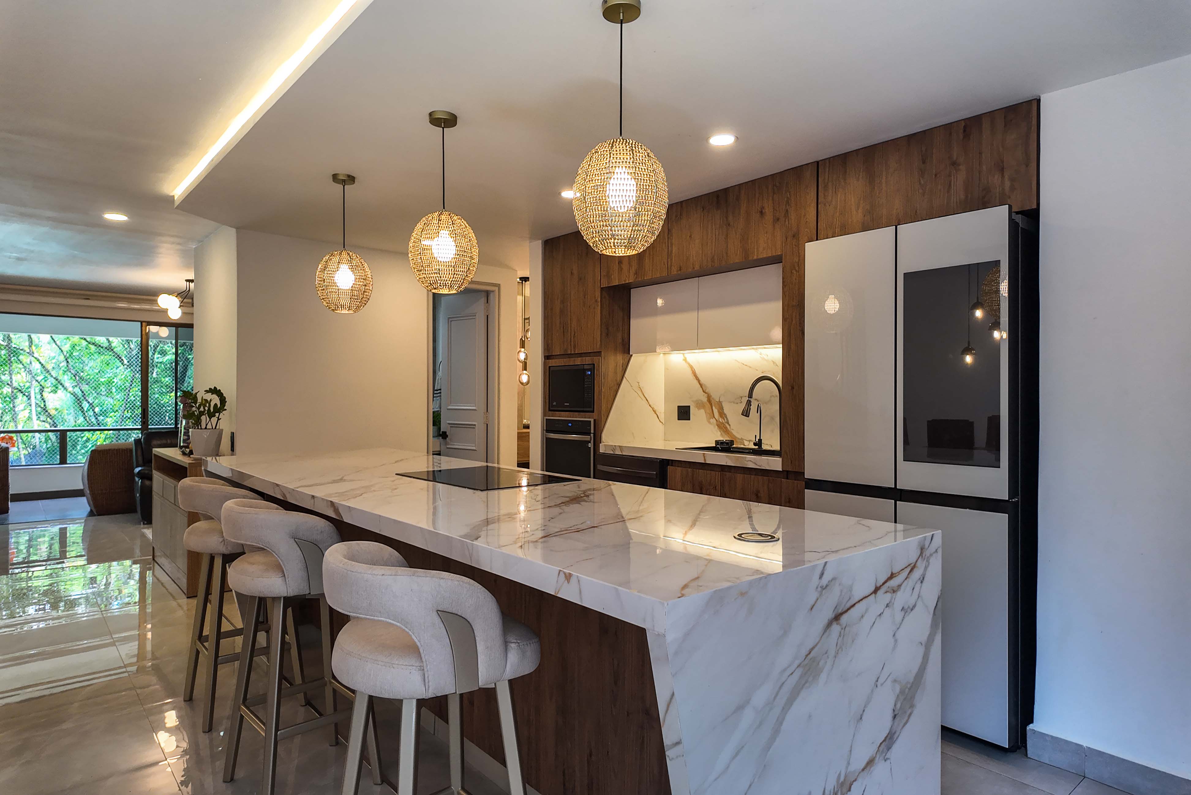 A modern kitchen with a marble island, beige stools, and woven pendant lights. Wood cabinets and a white refrigerator are in the background.
