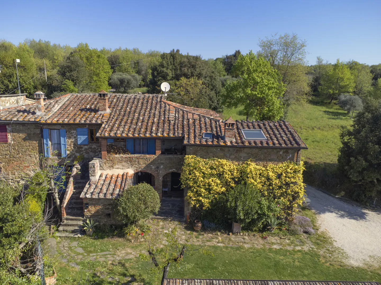 Exterior view of a stone house with a red tile roof, blue shutters, and yellow flowering vines. Green trees and grass surround the house.