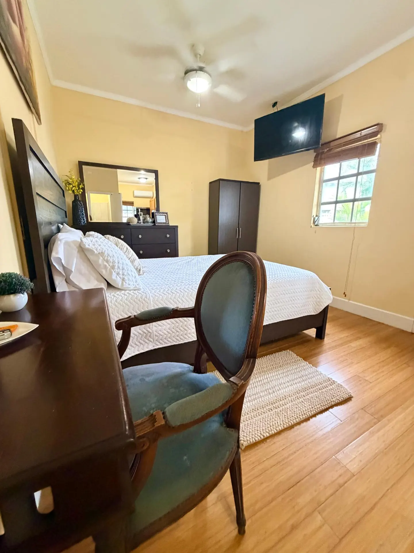 Bedroom with yellow walls, wood floors, and a bed with white linens. A blue chair sits near a desk. A TV is mounted on the wall.