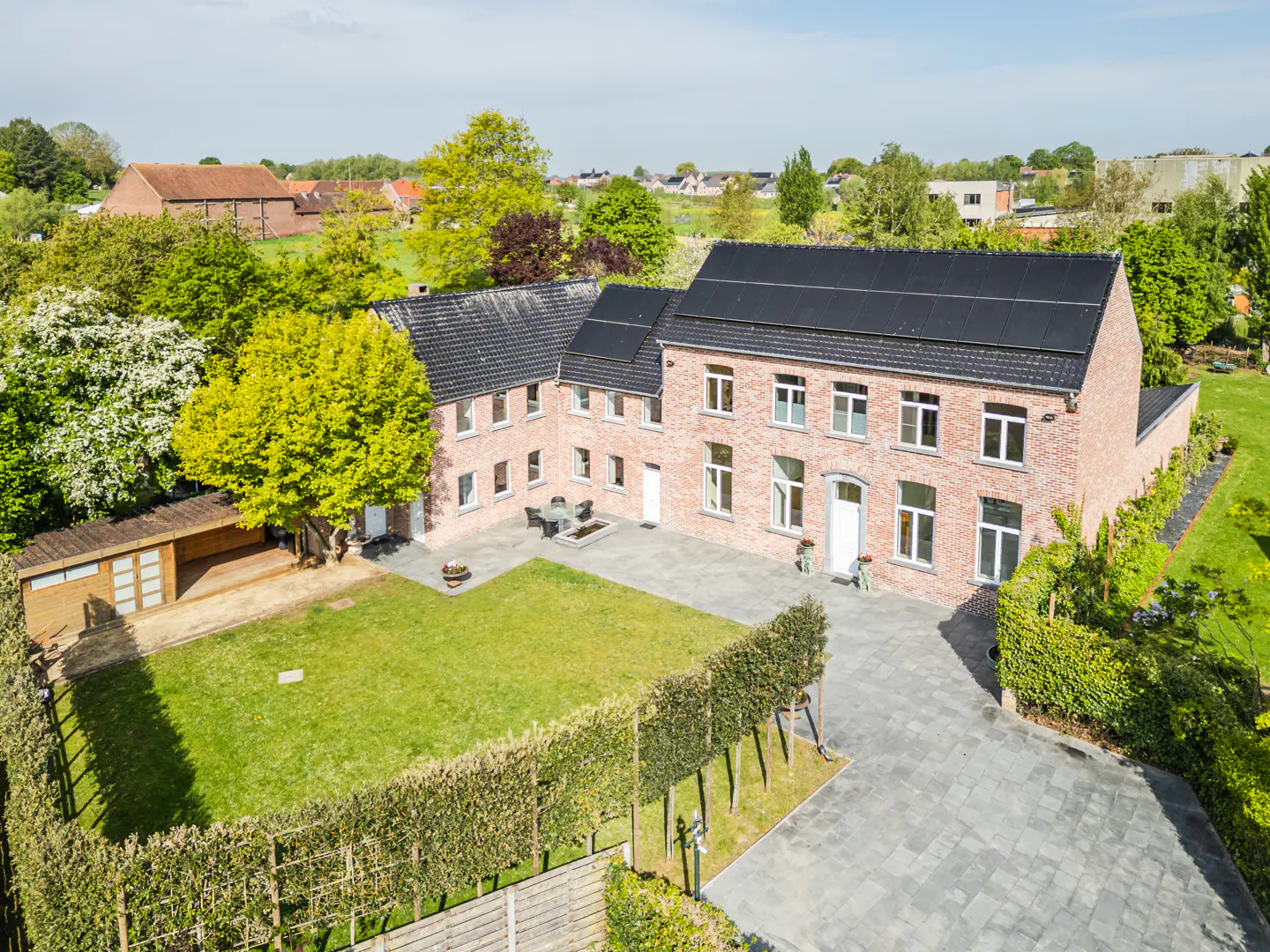 Aerial view of a two-story brick house with a black roof and solar panels, surrounded by a green lawn, trees, and a gray stone driveway.