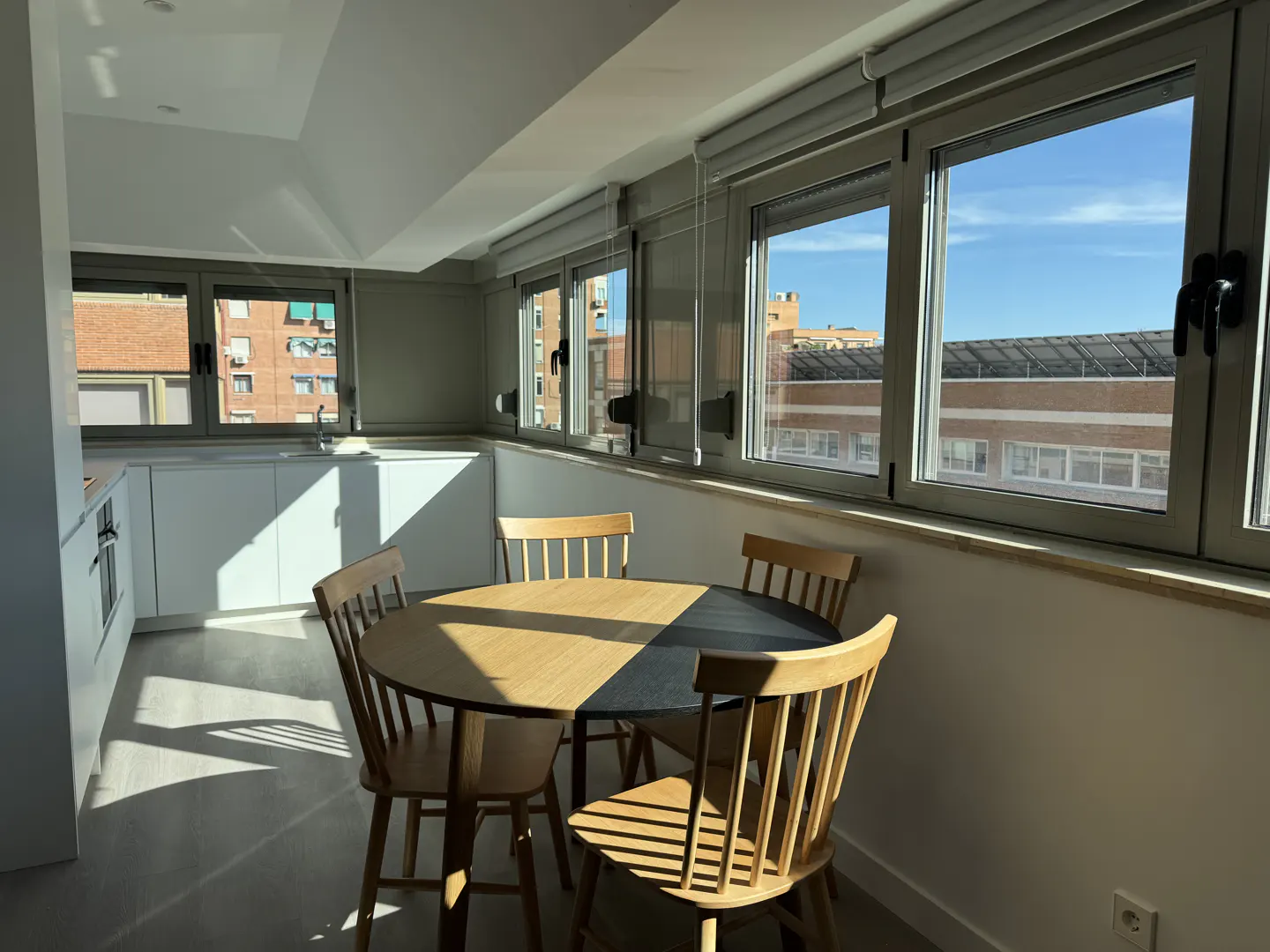 Bright kitchen with a round wooden table and four chairs, next to a row of windows with a view of buildings.