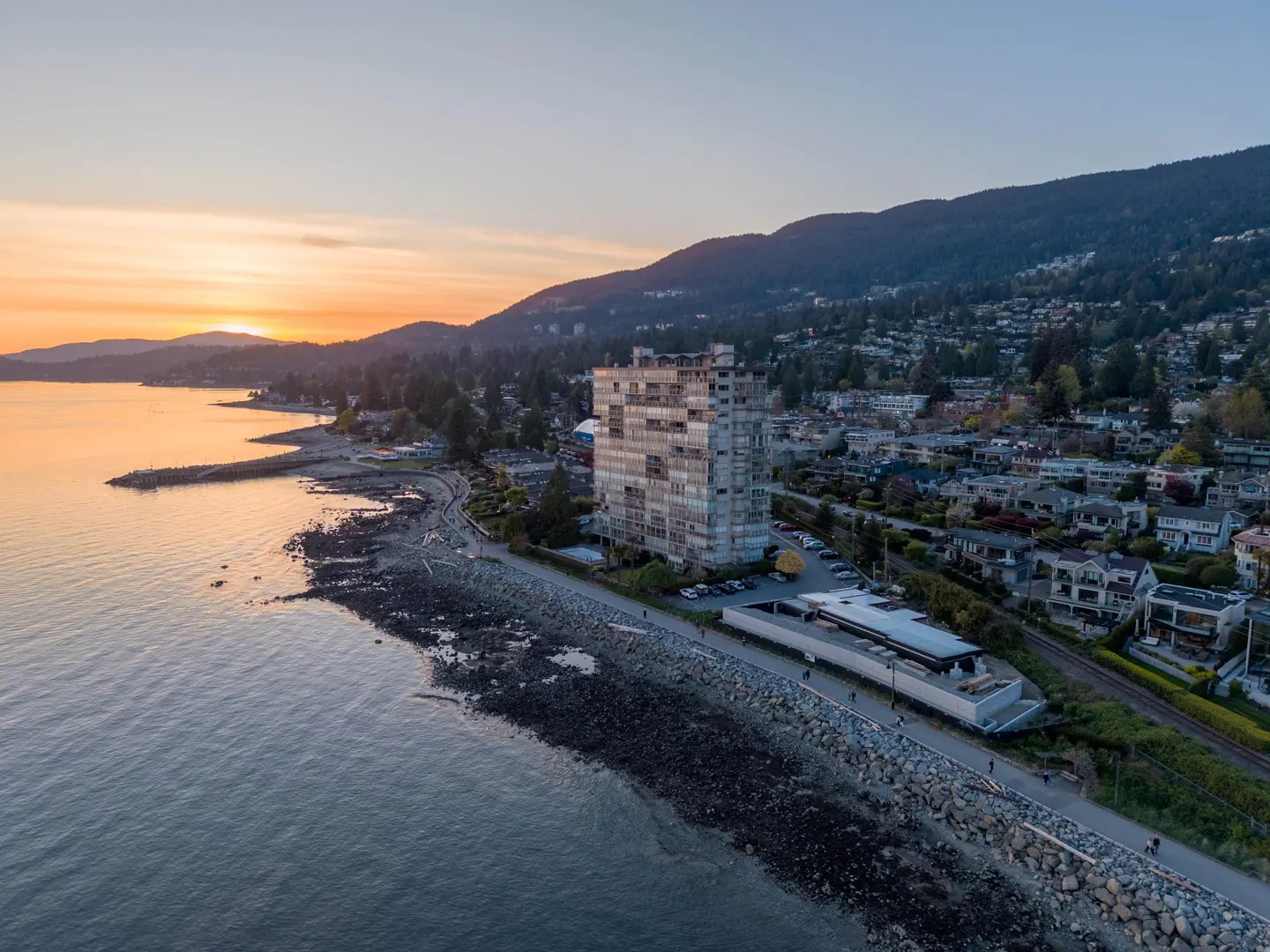 Aerial view of a tall building on a rocky beach at sunset, with mountains and houses in the background.