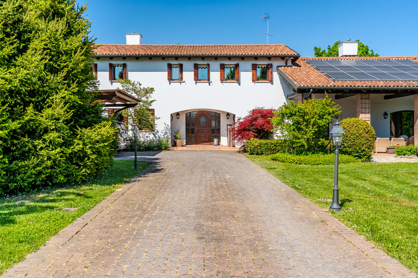 Exterior view of a two-story white house with a red tile roof and solar panels, seen from a paved driveway.