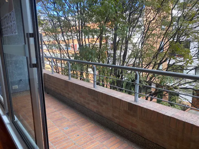 Balcony view with brick flooring, a low brick wall, and a metal railing. Trees and buildings are visible in the background.