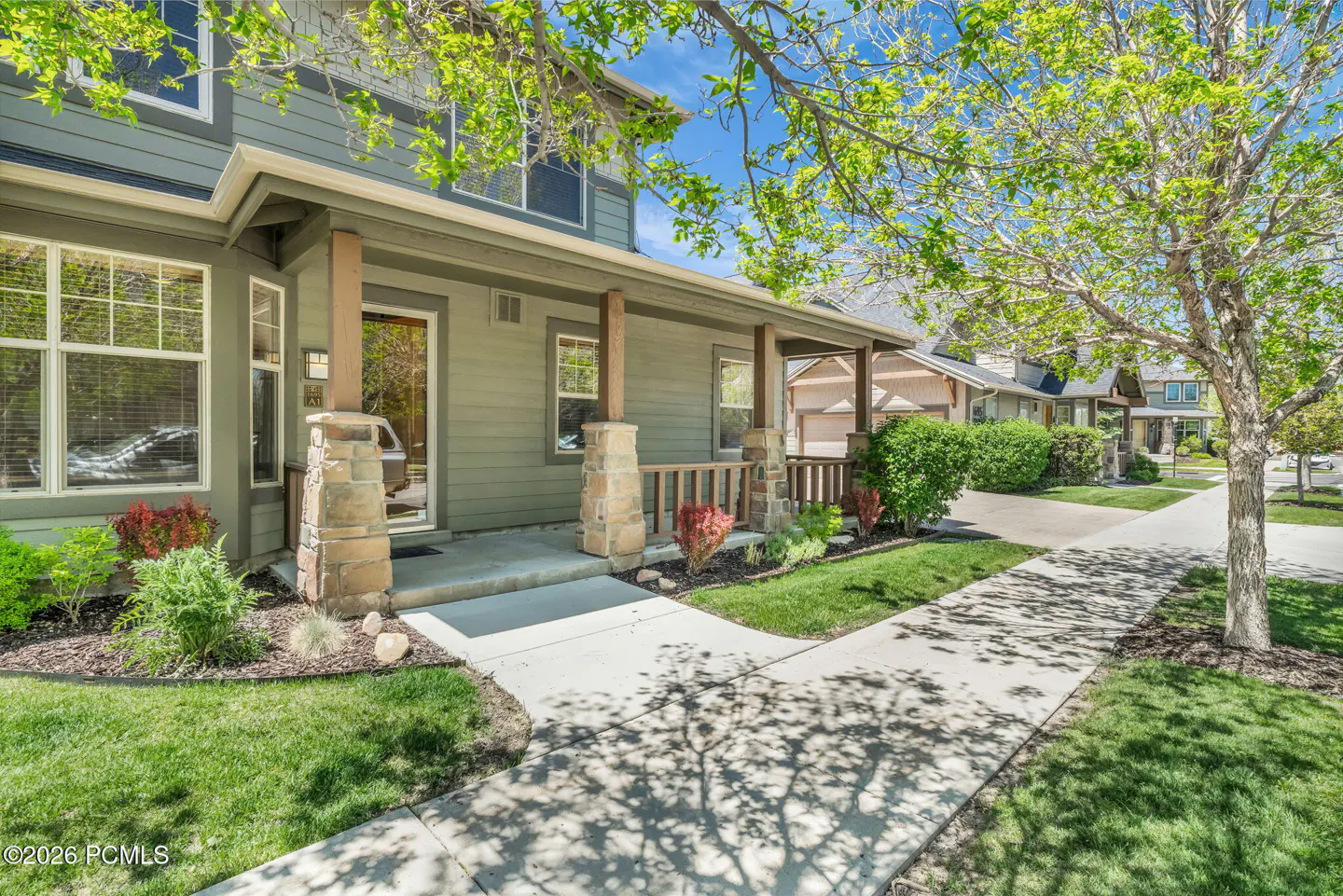 Green two-story house with a stone-pillared porch, sidewalk, and leafy trees under a bright blue sky.