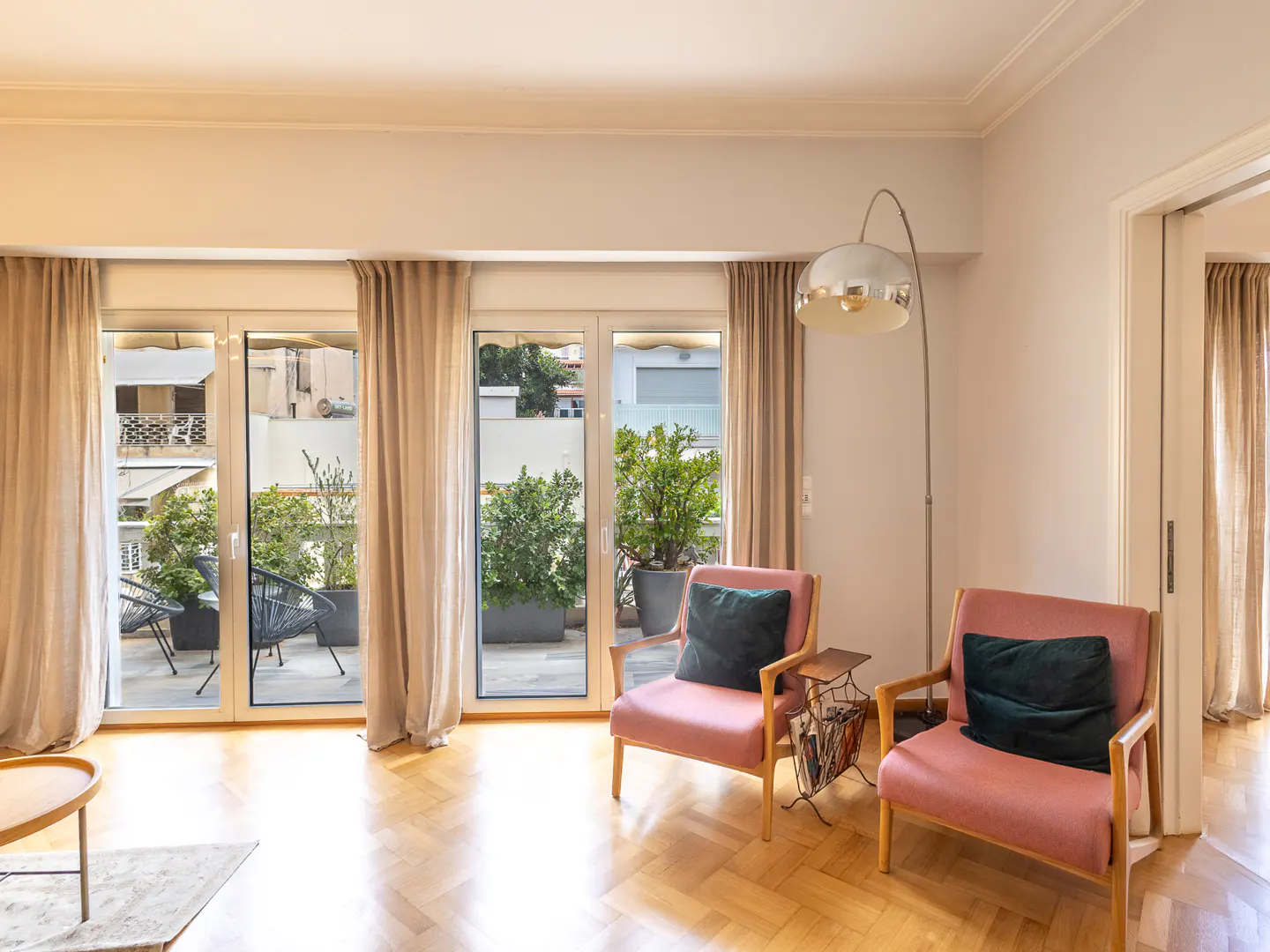 Two pink armchairs with dark cushions sit on a herringbone wood floor near a floor lamp and glass doors to a balcony.