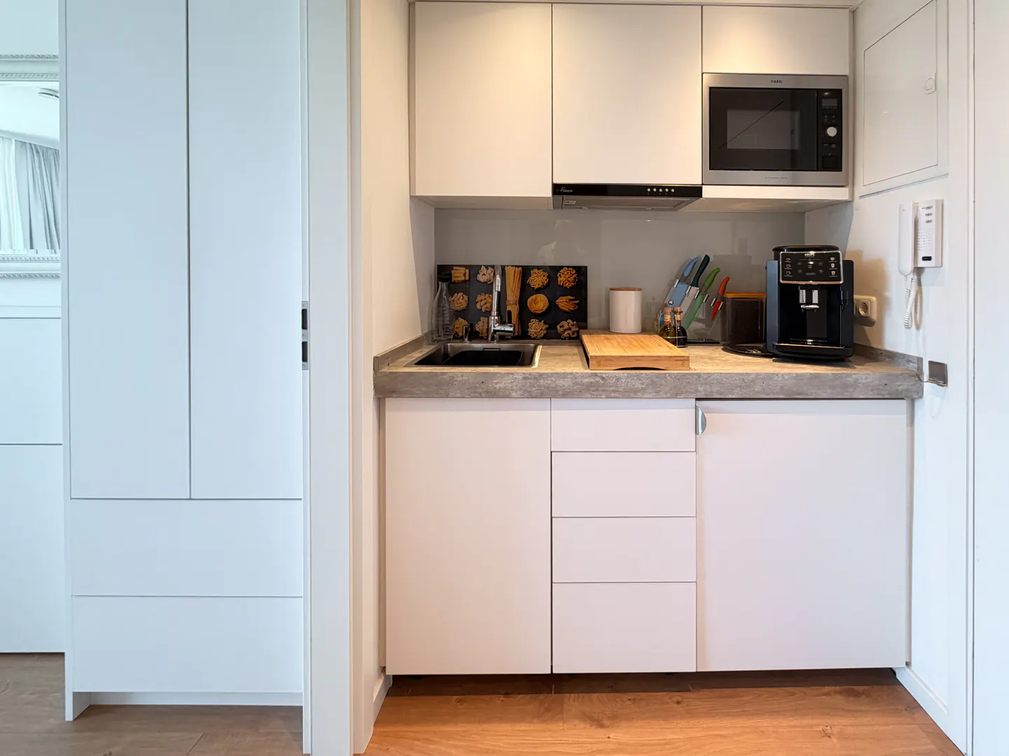 Modern kitchenette with white cabinets, grey stone countertop, built-in microwave, coffee machine, and wood flooring.