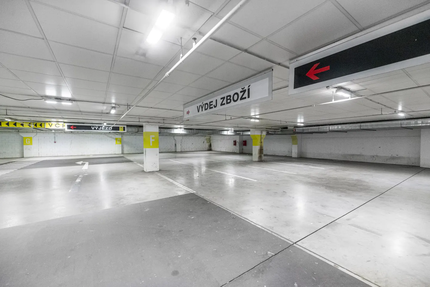 Indoor parking garage with gray concrete floor, white ceiling, and yellow support columns. Directional signs hang from the ceiling.