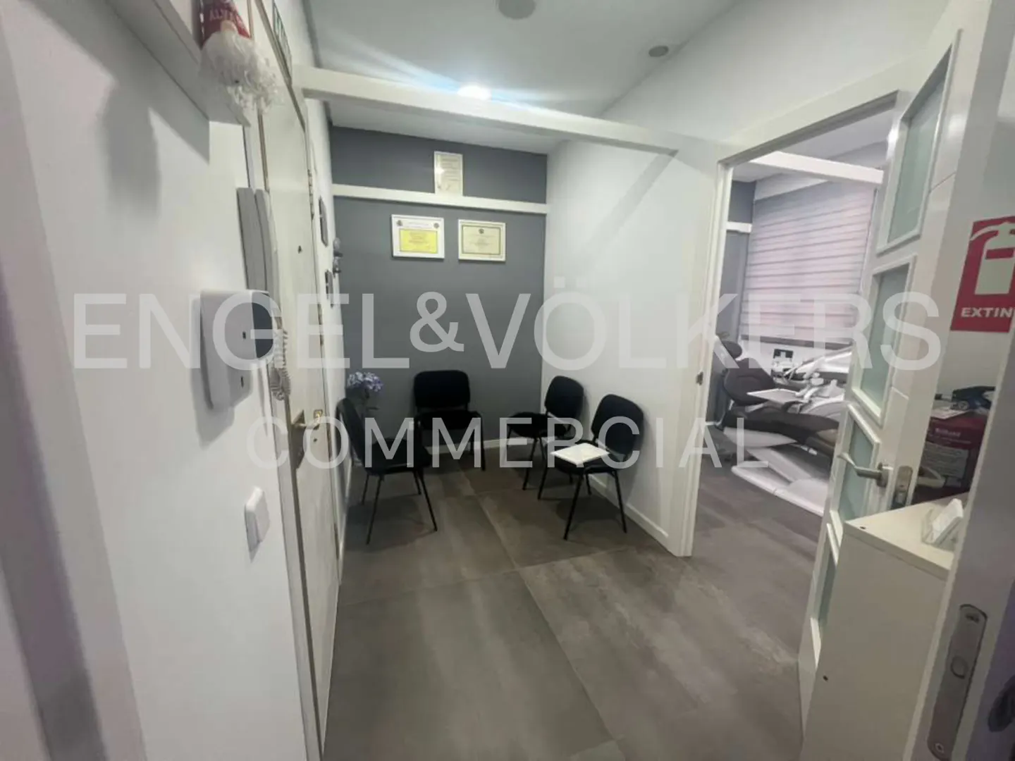 Waiting room with black chairs, gray walls, and certificates. A dental chair is visible through an open door. Engel & Volkers logo overlay.