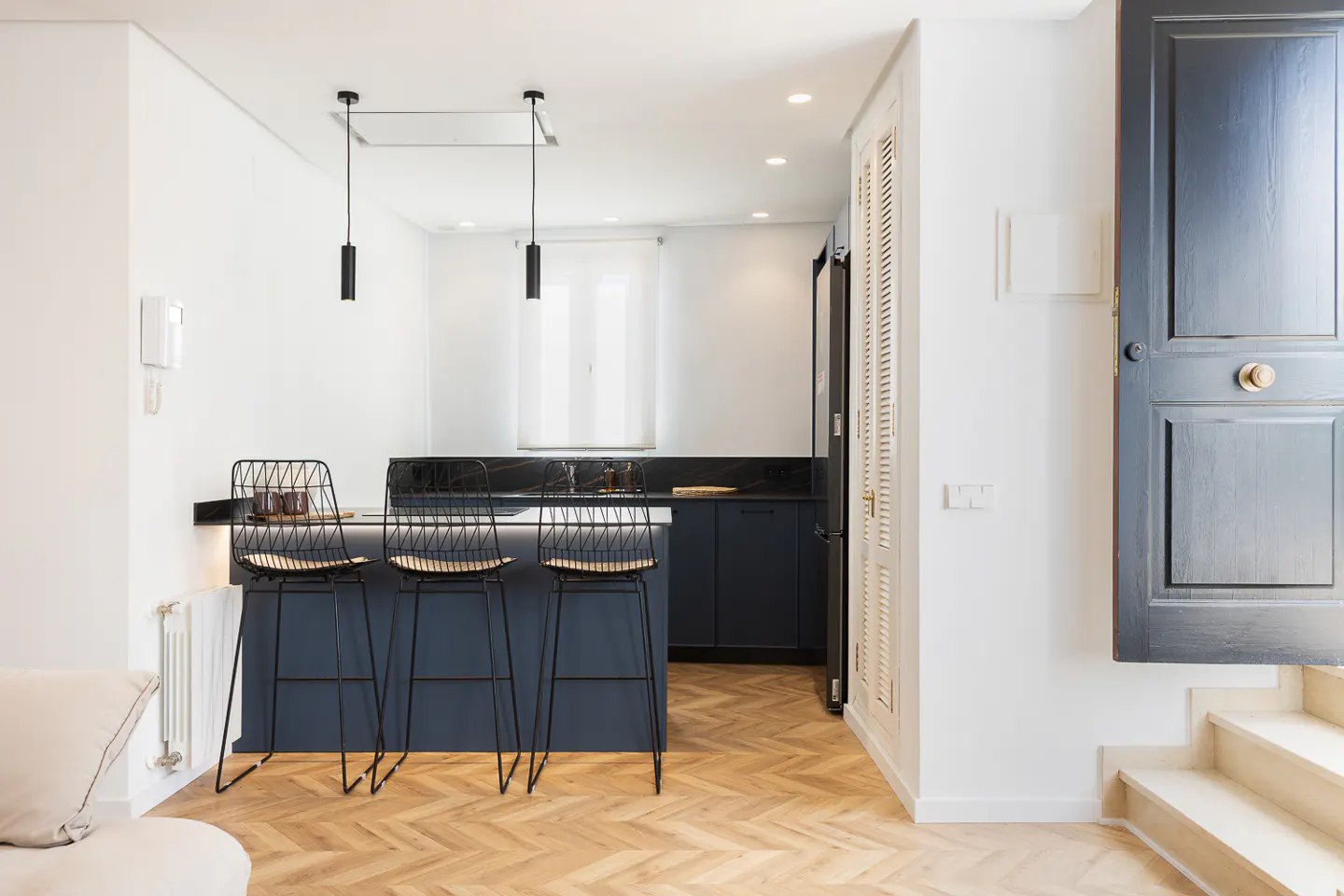 A modern kitchen with a blue island, black wire stools, and herringbone wood floors. A black door is partially open on the right.