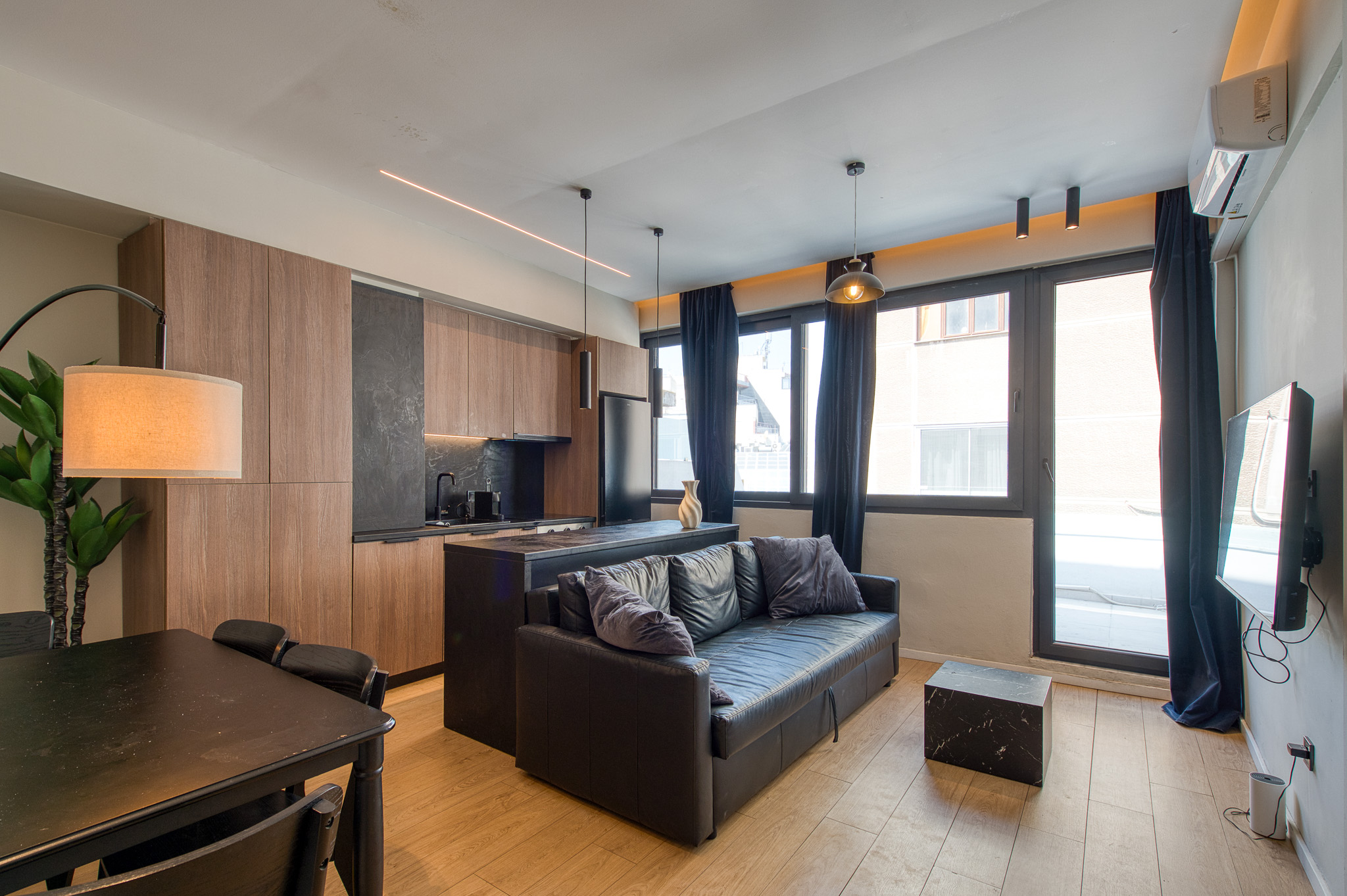 Modern studio apartment with a black sofa, kitchen island, wood cabinets, dining table, and large windows with dark curtains. [AI-generated text]