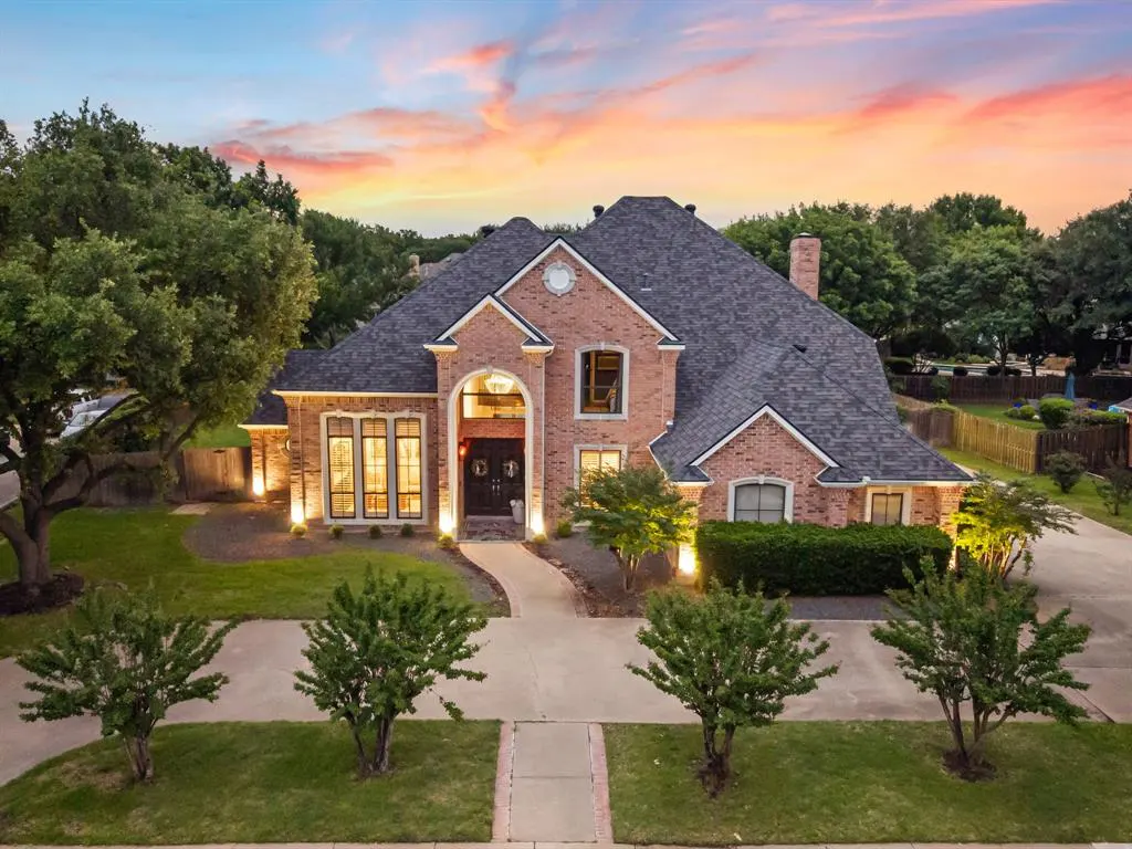 Two-story brick house with a gray roof, green lawn, and trees under a colorful sunset sky.