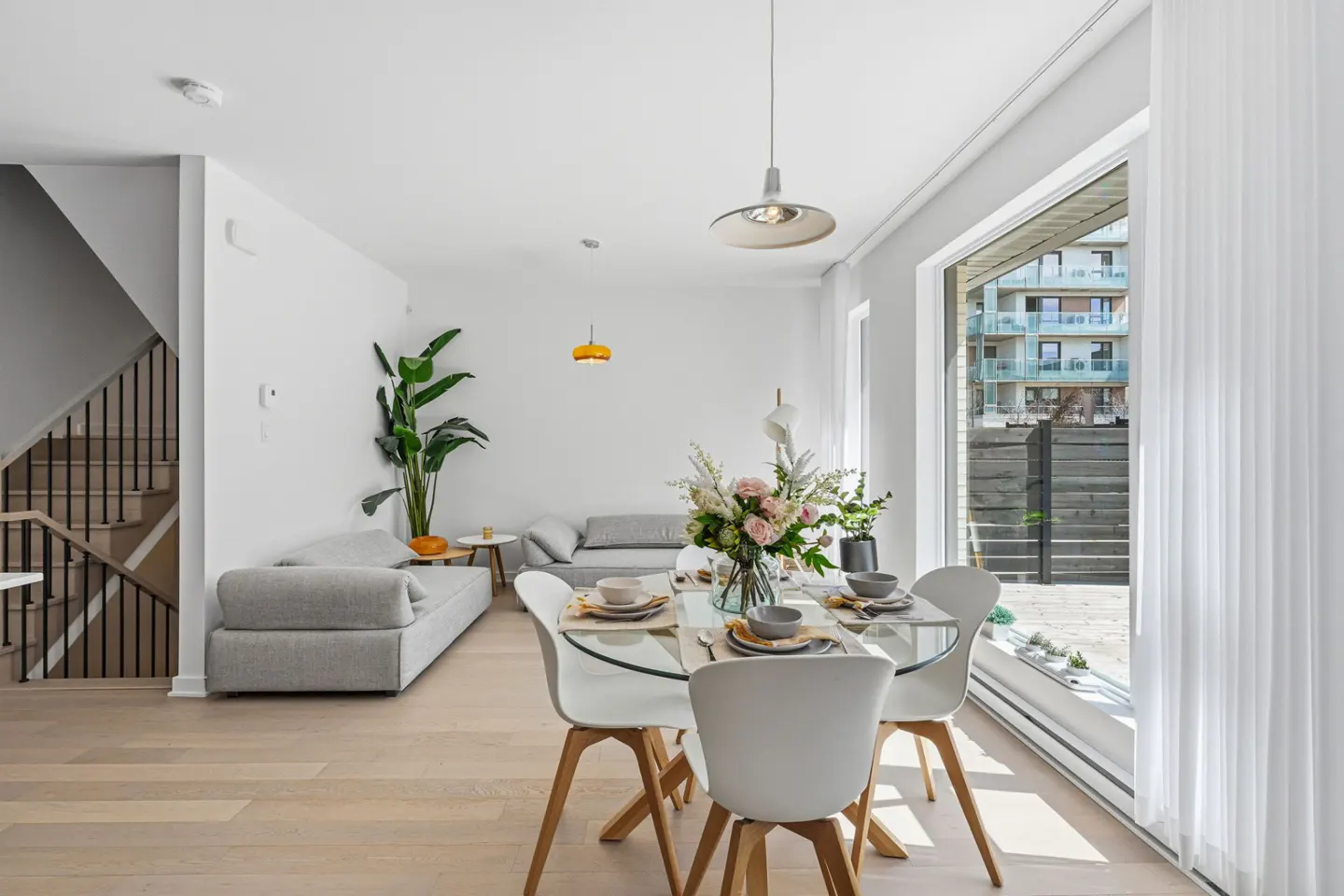 Bright, modern living space with a glass dining table, white chairs, and a gray sofa. Large window with sheer curtains and a view of an apartment building.