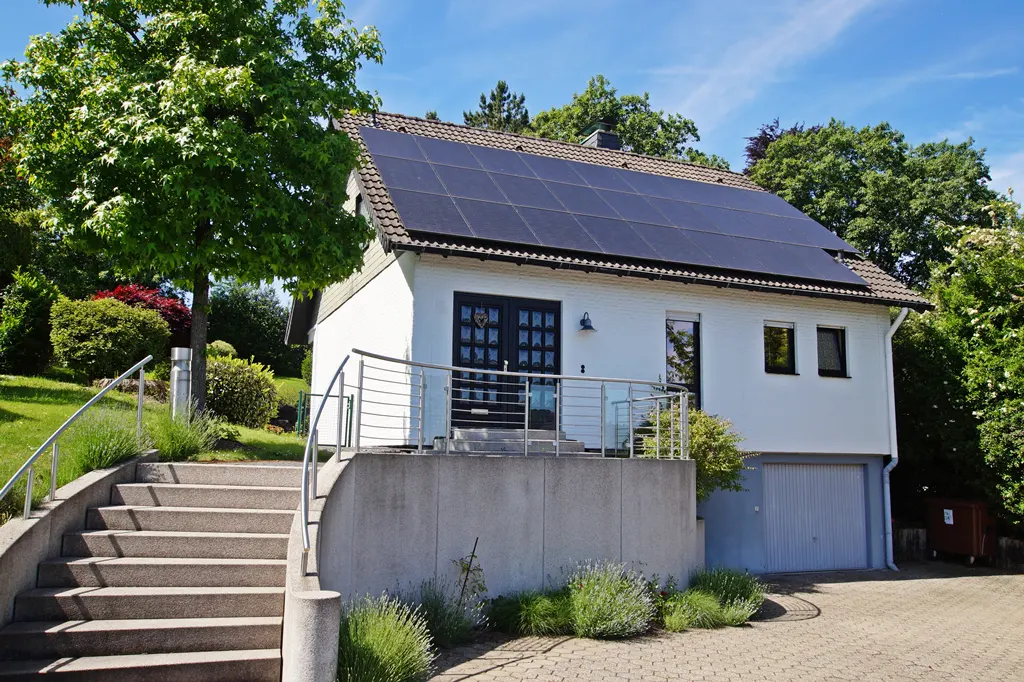 White house with solar panels on the roof, a front staircase with metal railings, and a garage on a sunny day.