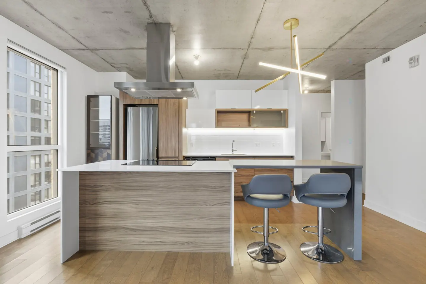 A modern kitchen with wood floors, a concrete ceiling, and a large window. Two gray bar stools sit at the island.