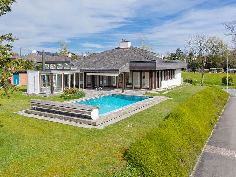 Exterior view of a modern house with a pool, green lawn, and trees under a blue sky.