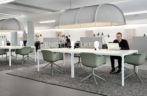 Modern office space with white desks, olive green chairs, and large gray sound dampening lights overhead. A man works on a laptop.
