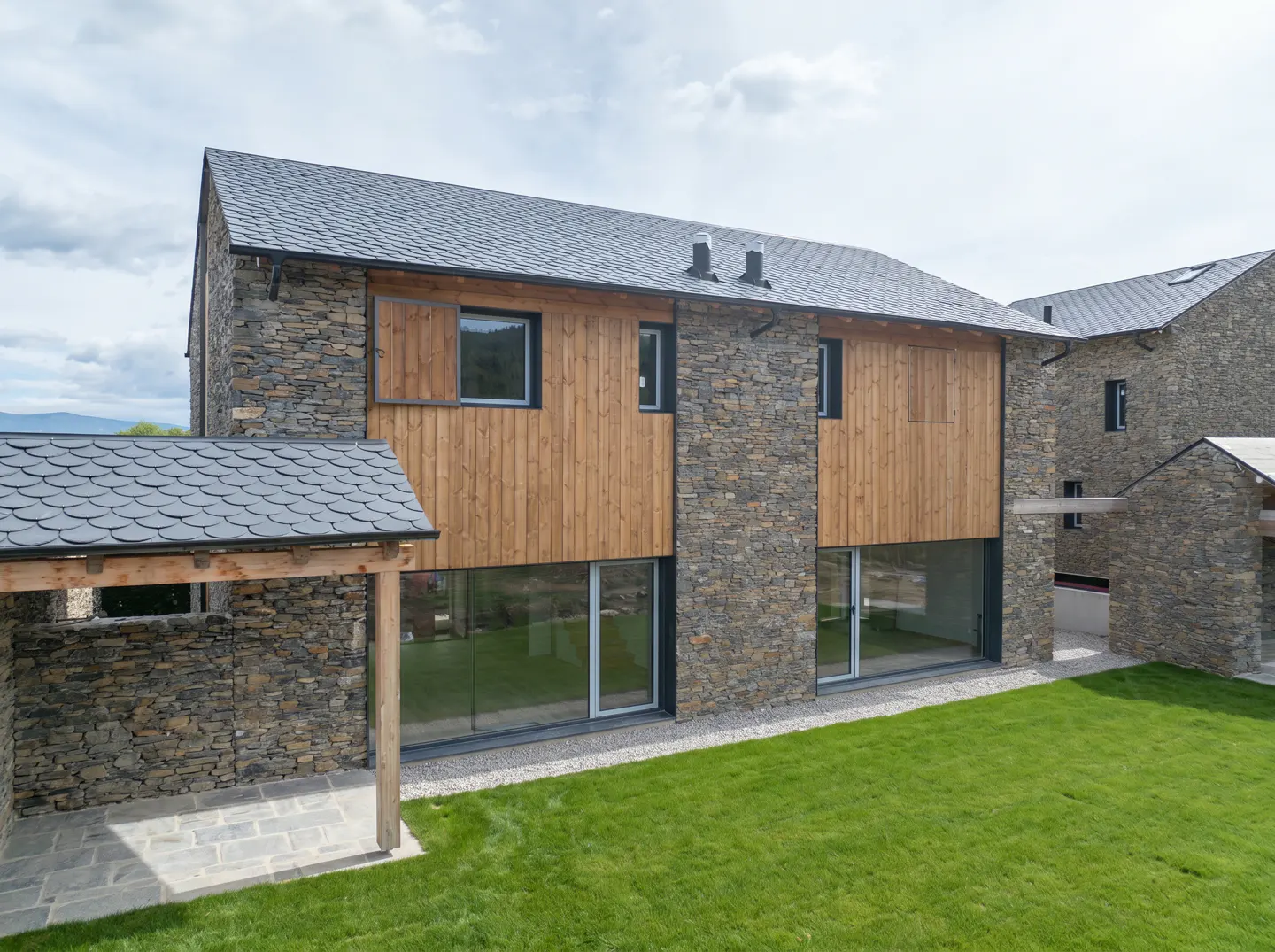 Modern house exterior with stone and wood facade, large windows, and a green lawn. Slate roof and covered patio visible.