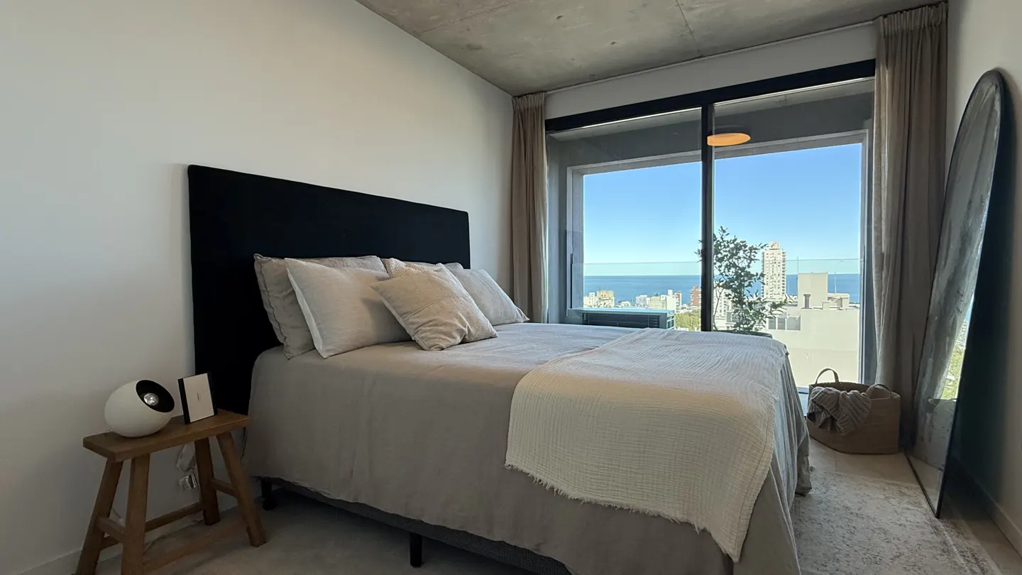 Bedroom with a black headboard, beige bedding, and a large window with a city and ocean view. A wooden stool and mirror are also visible.
