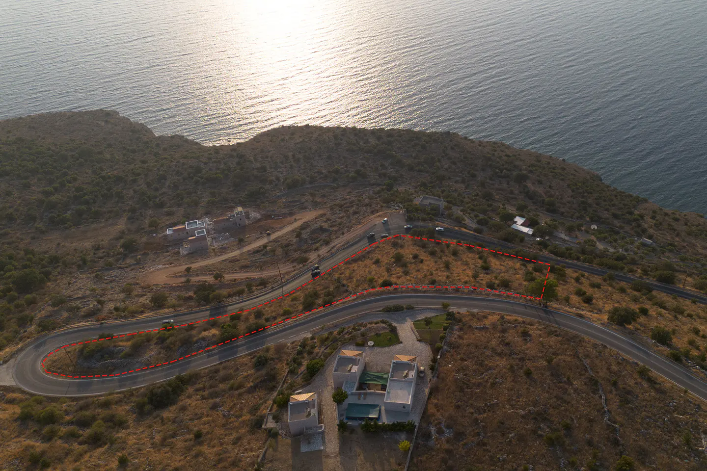 Aerial view of a coastal hillside plot outlined in red, situated along a winding road overlooking the sea.