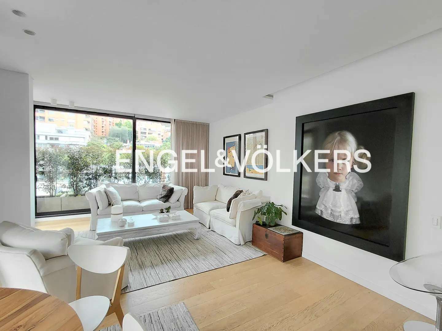 Bright living room with white sofas, a white coffee table, and a large framed portrait on the wall. A wooden chest sits near a potted plant.