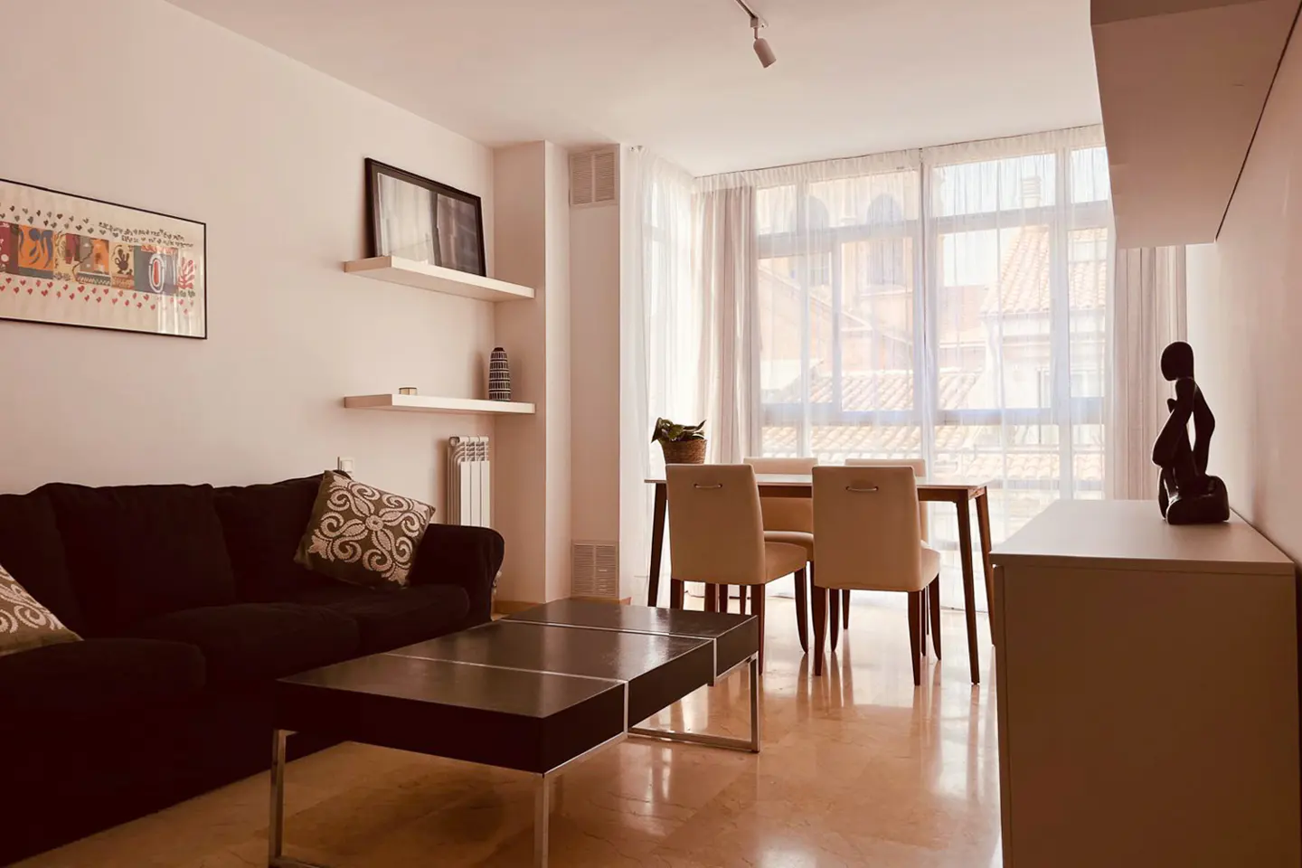 Living room with a black sofa, coffee table, dining table with chairs, and white shelves with decor. Large windows with sheer curtains.