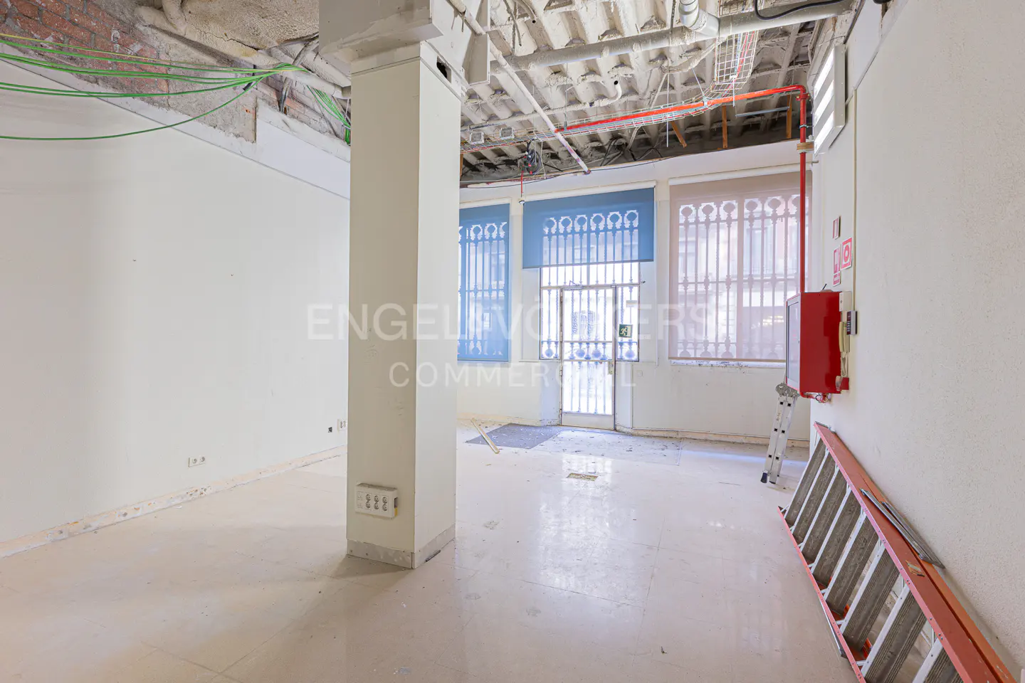 Unfinished commercial space with white walls, exposed ceiling, and blue window shades. A red fire hose box and ladder are on the right wall.