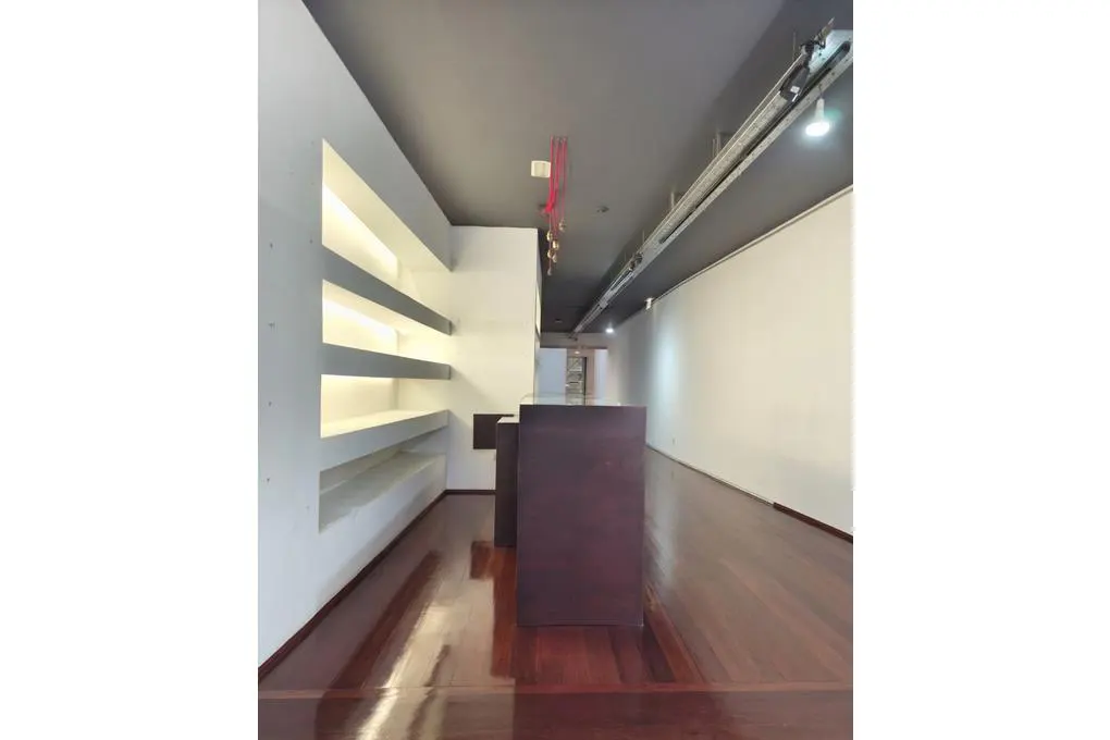 Empty retail space with wood floors, white walls, and gray ceiling. Built-in shelves with lighting on the left, and a dark brown counter in the center.