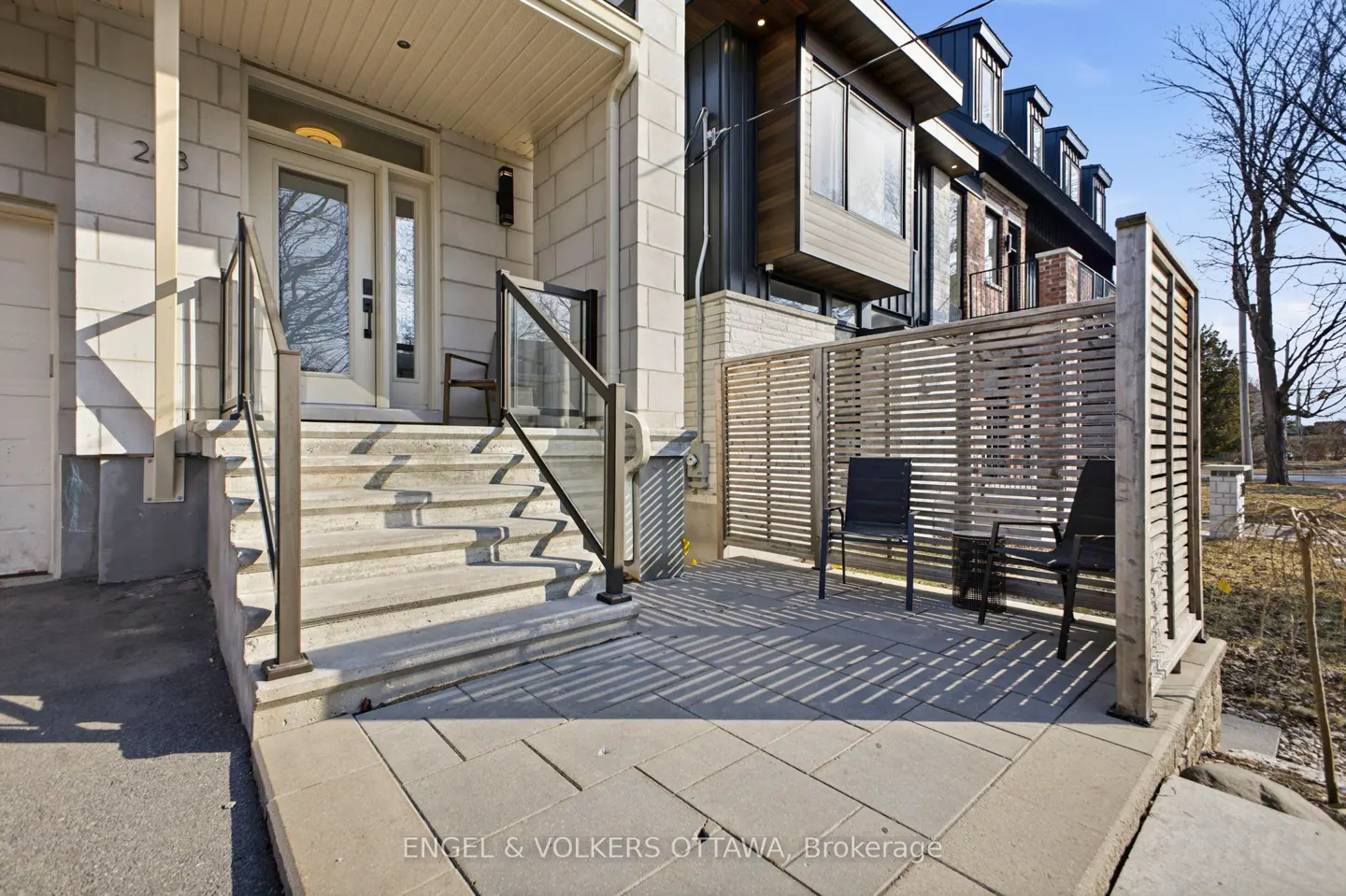 Exterior view of a modern home with stone steps leading to a white front door. A patio with a wooden screen and two black chairs is visible.