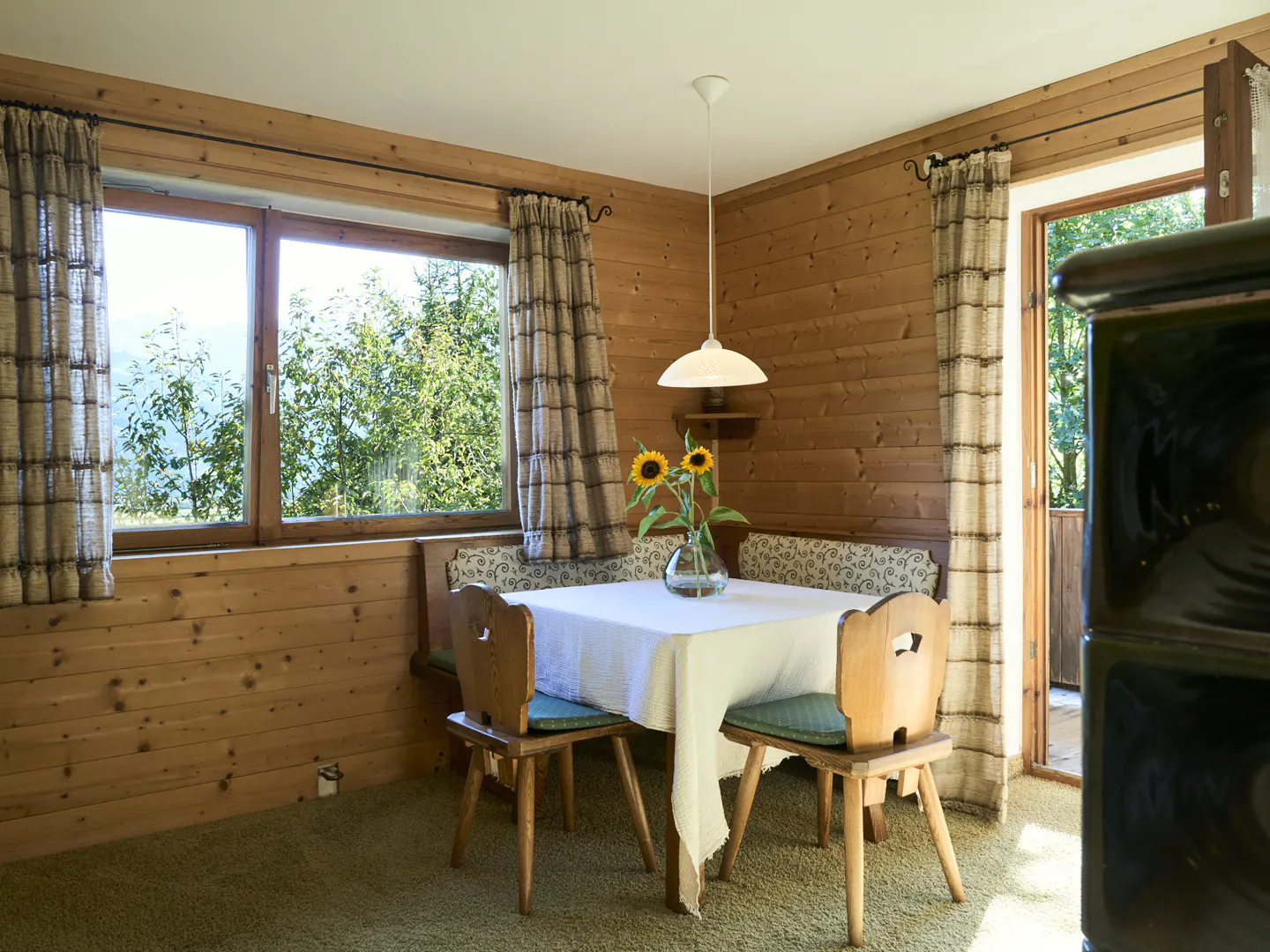 Cozy dining area with wood-paneled walls, a window with a view, and a table set with sunflowers. Curtains frame the window and doorway.