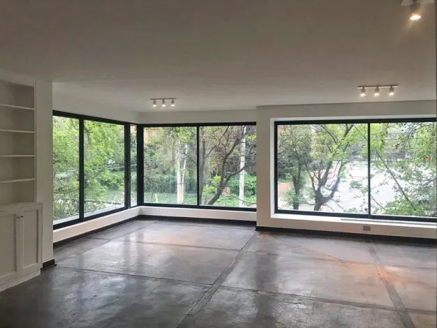 Empty room with gray concrete floors, white walls, and large windows with black frames overlooking green trees.