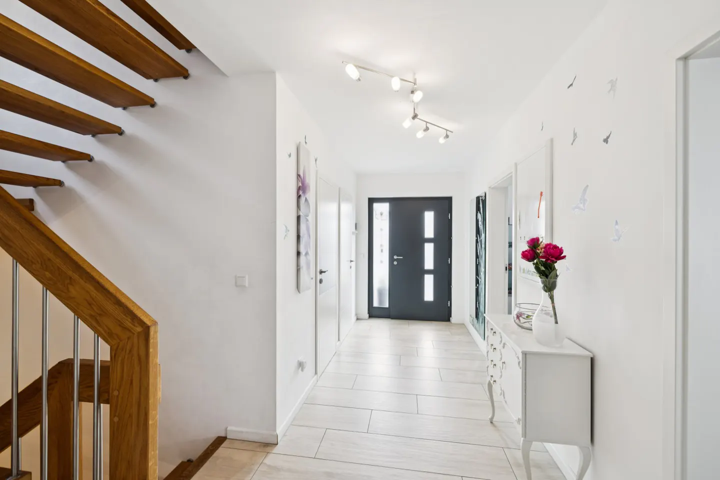 Bright hallway with white walls, light tile floor, and a dark front door. A wooden staircase is on the left, and a white console table with flowers is on the right.