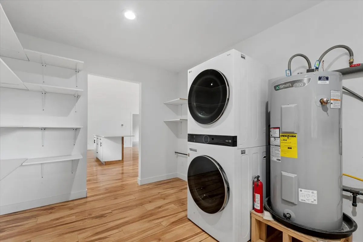 Bright laundry room with wood floors, white walls, and open shelving. Stacked white washer/dryer next to a gray water heater.