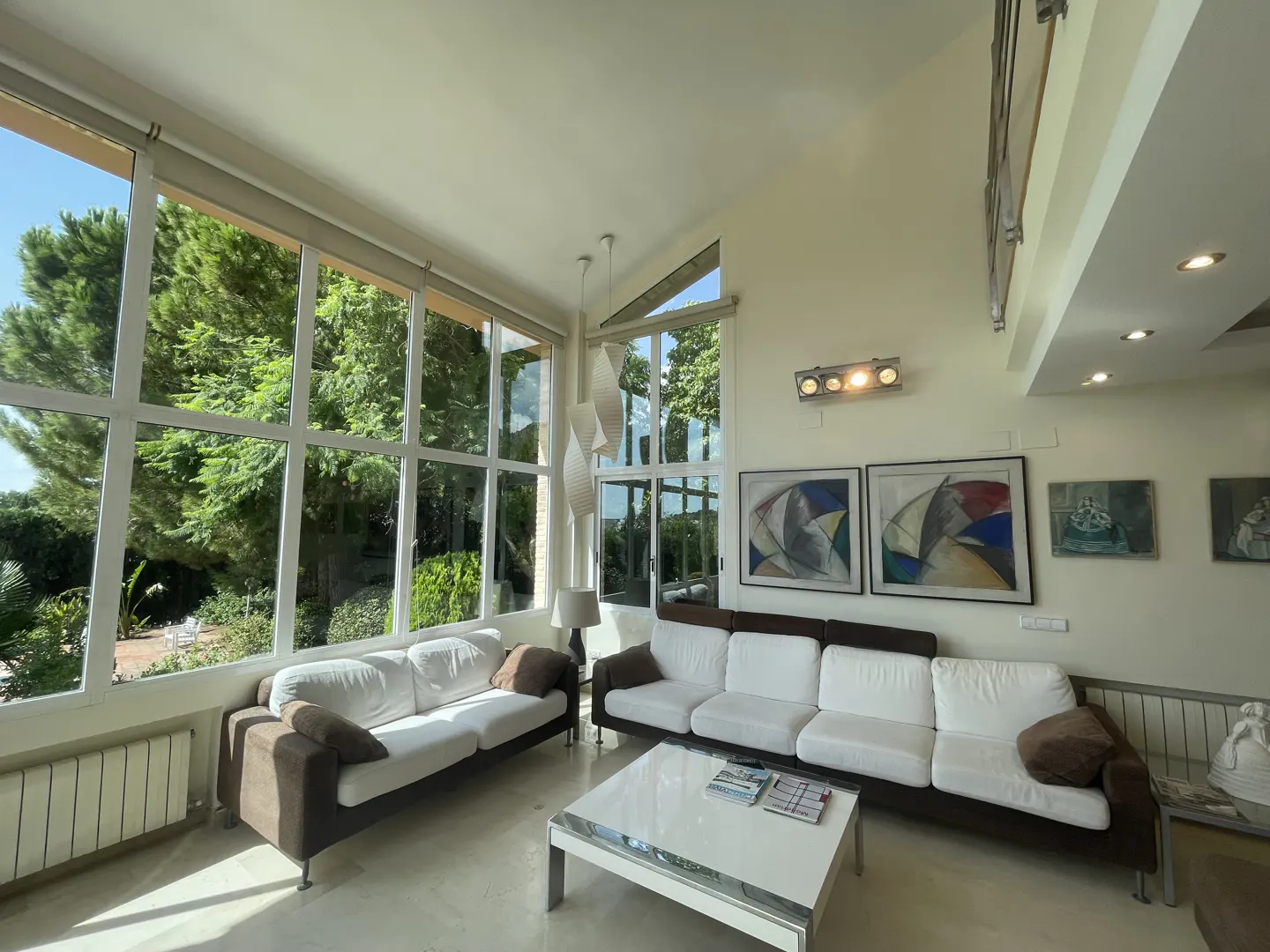 Bright living room with white sofas, brown accents, and large windows overlooking lush greenery. Modern art adorns the walls.