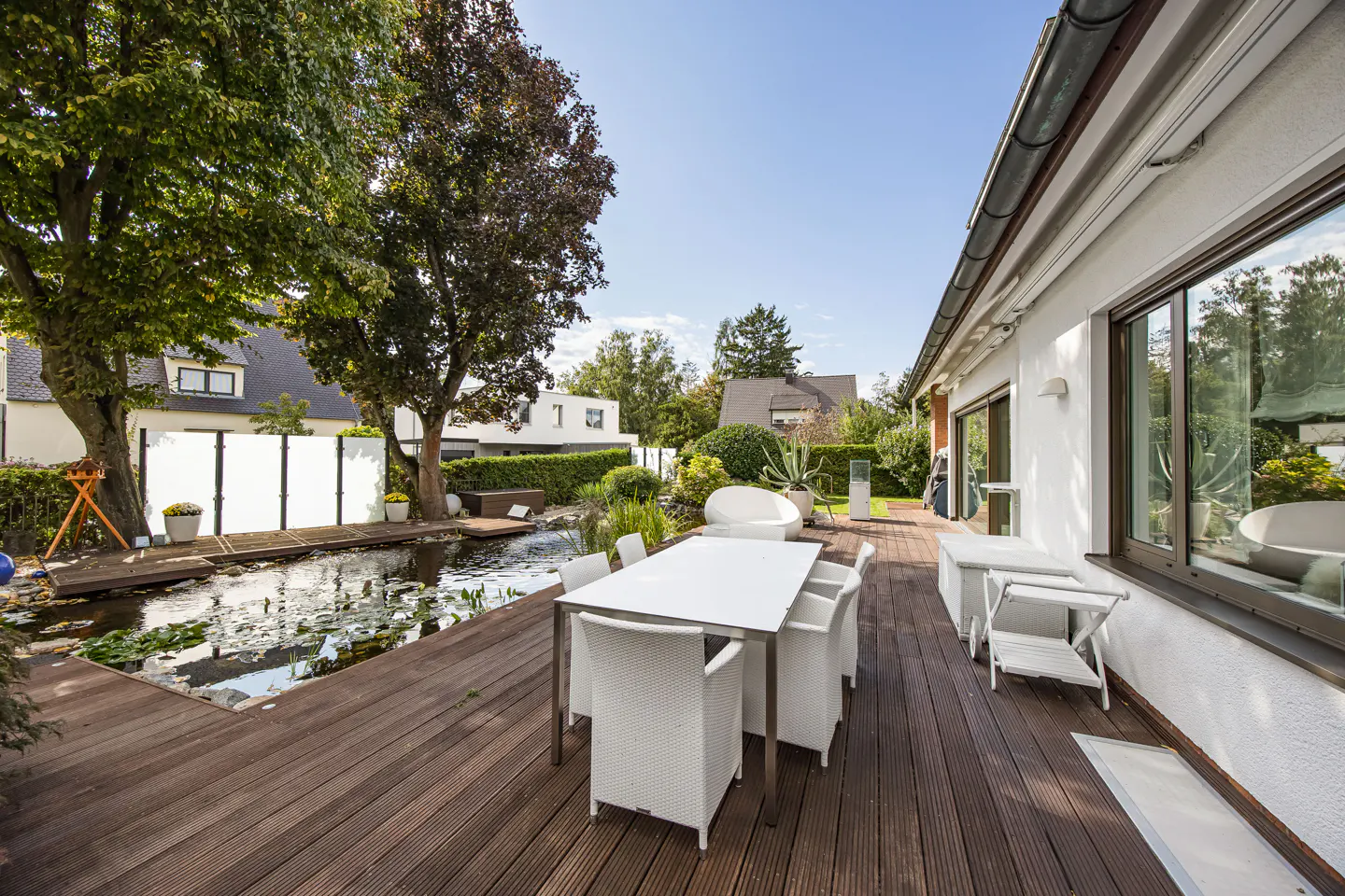 A backyard patio with a pond, a white table and chairs on a wooden deck, and a white house.