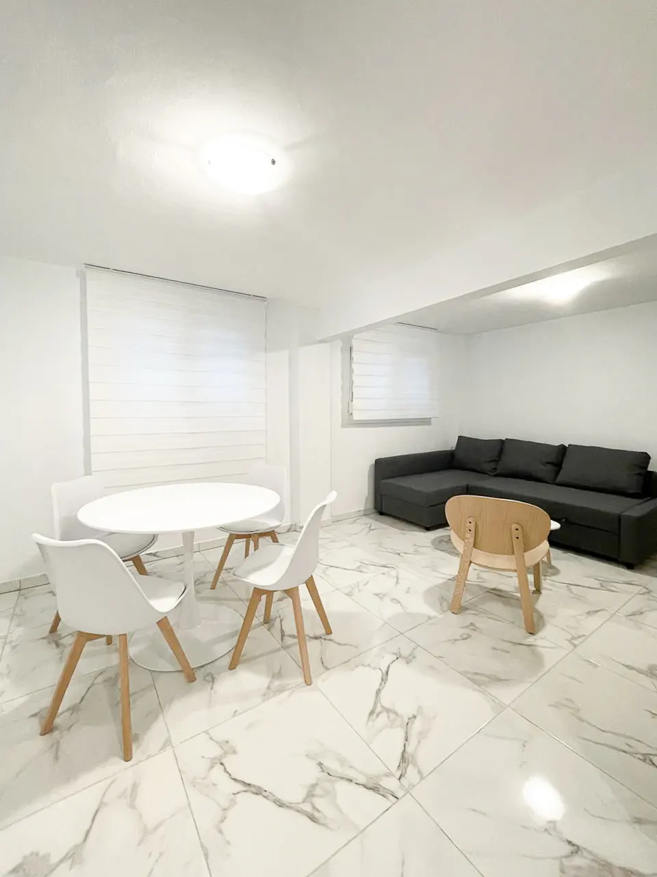 Bright room with white walls, marble floors, and two seating areas. A white table with chairs and a gray sectional sofa with a wooden coffee table.
