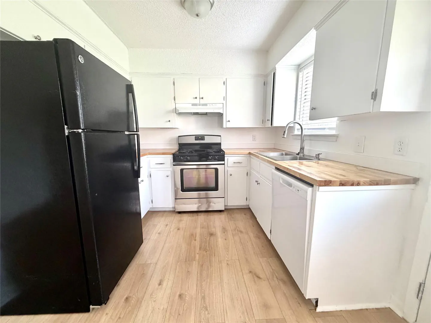 A bright kitchen with white cabinets, wood countertops, stainless steel appliances, and a black refrigerator.
