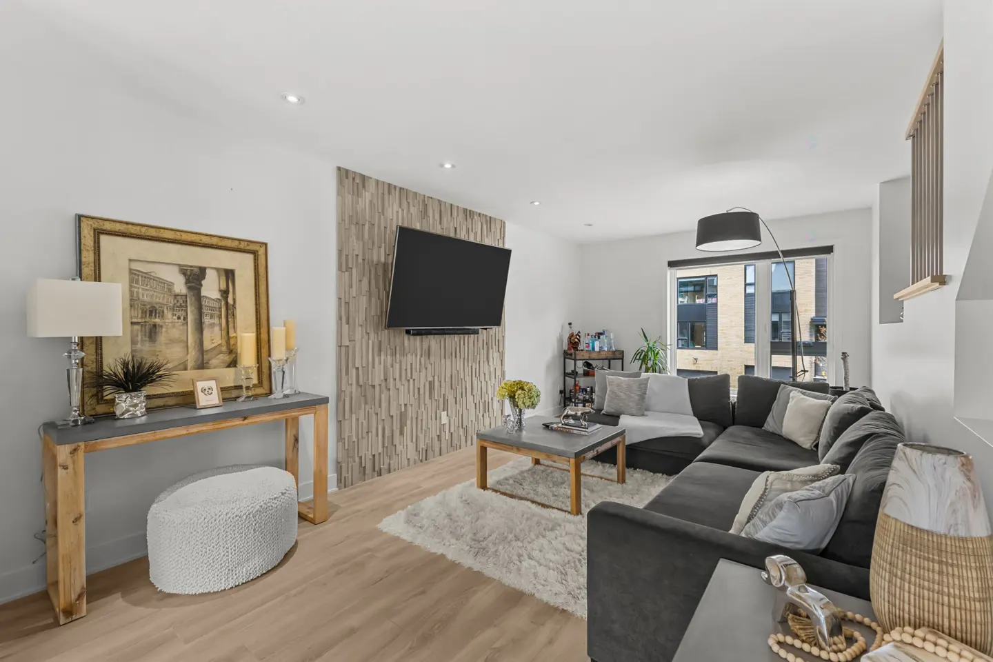 Bright living room with a gray sectional sofa, a TV mounted on a stone accent wall, and a console table with a lamp.