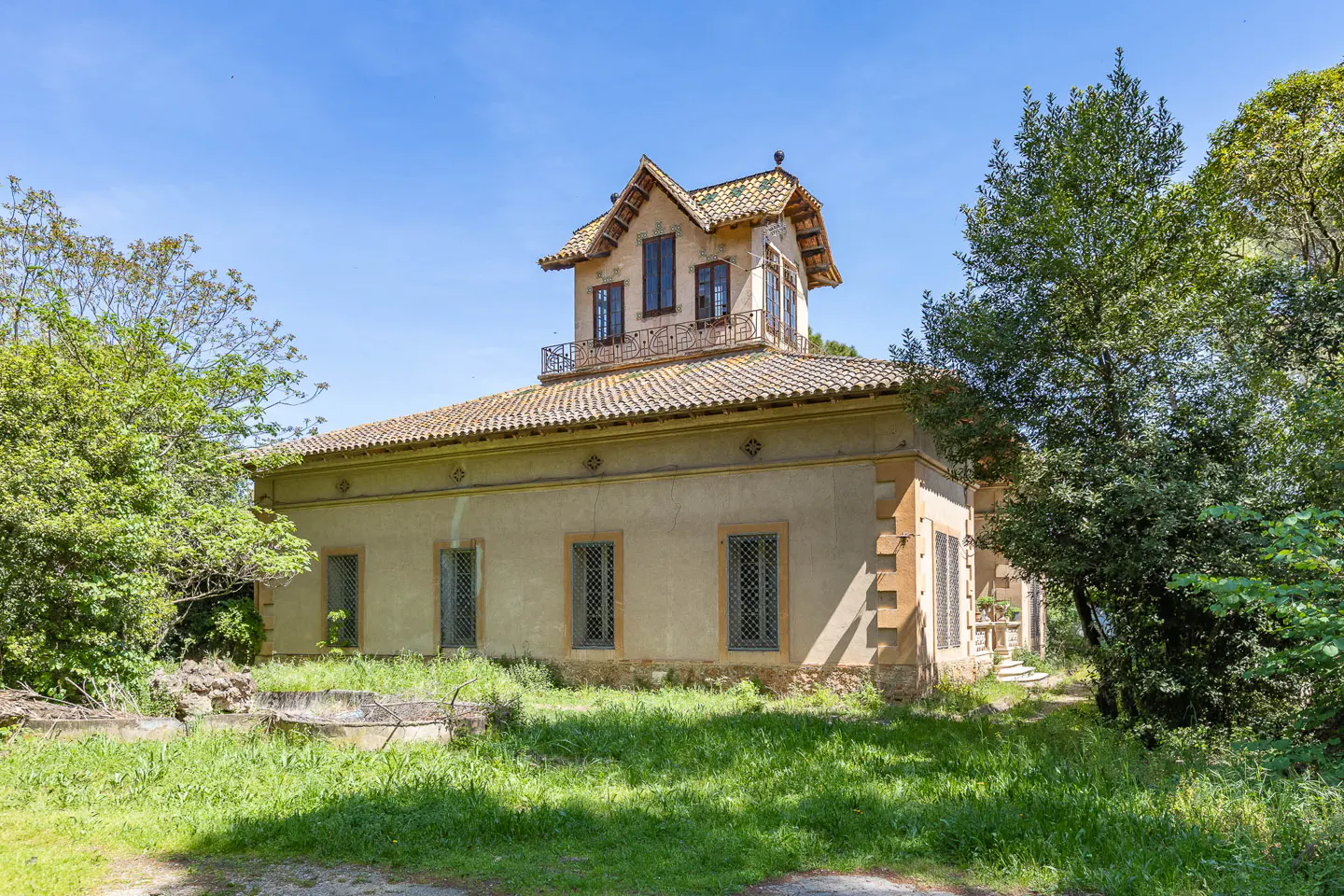 Exterior view of a beige, two-story house with a tiled roof and a small balcony, surrounded by green trees and grass.