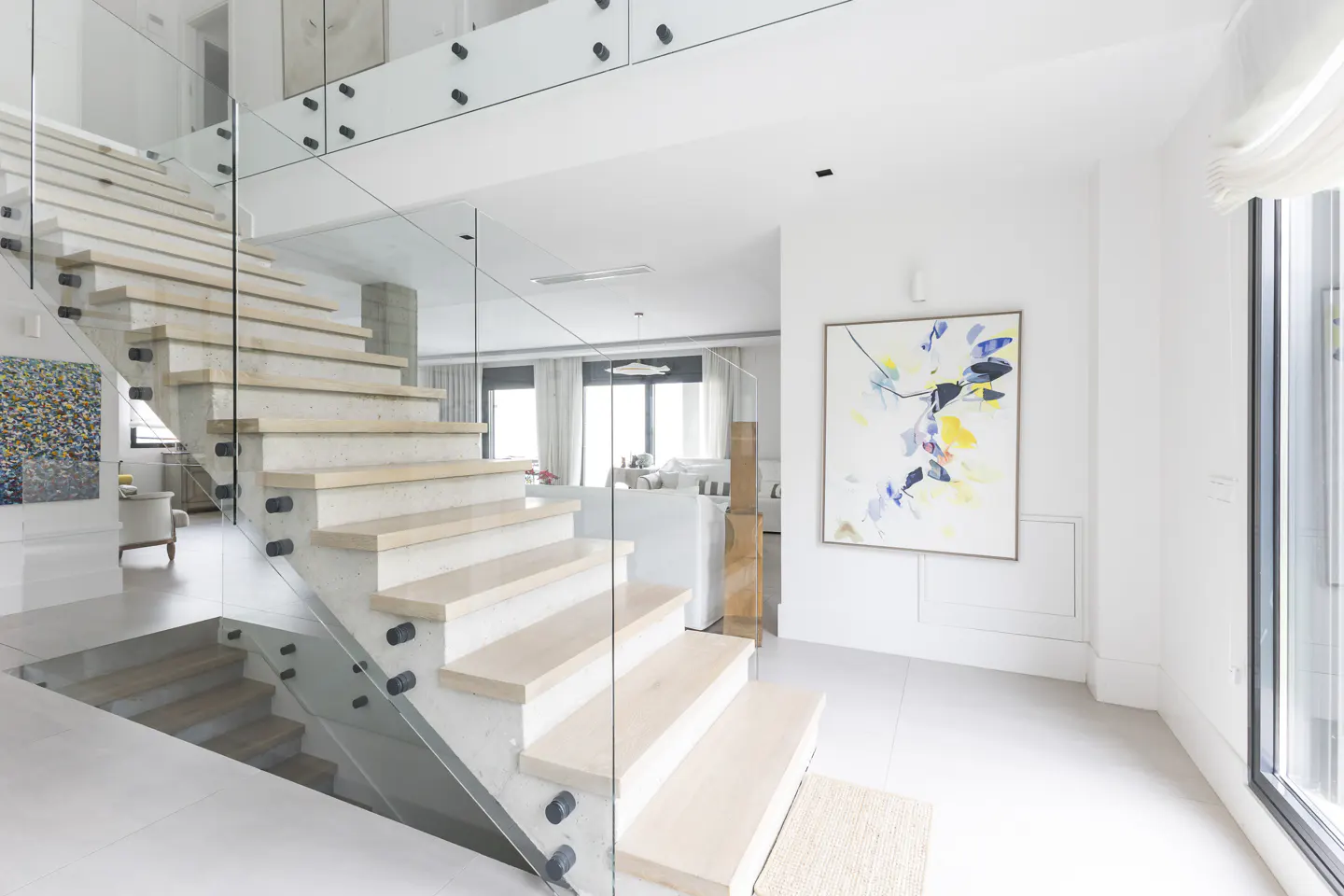 Bright, modern foyer with light wood stairs, glass railings, and white walls. An abstract painting hangs on the wall.